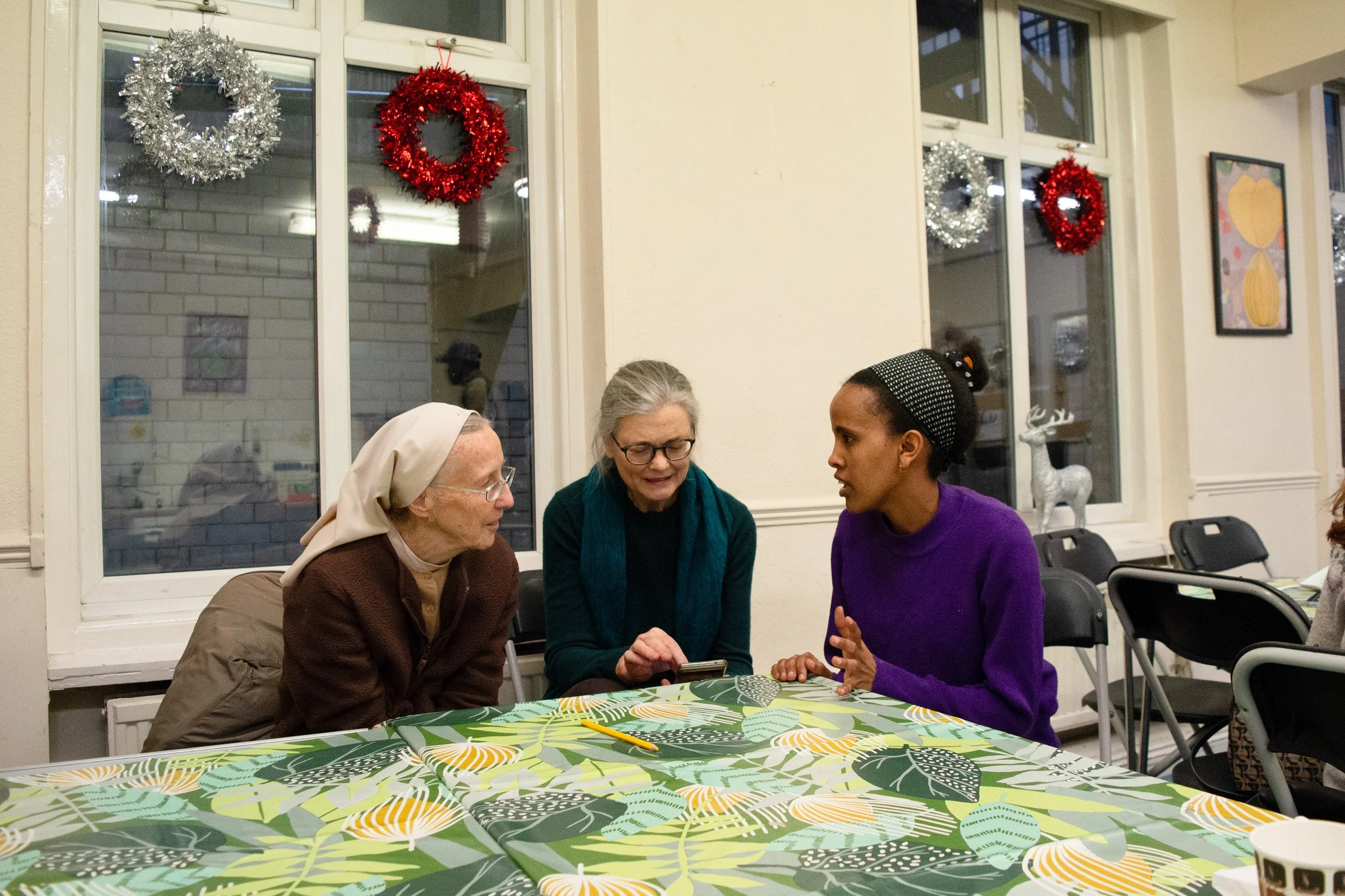 Three women are sitting around a table with a green leafy patterned tablecloth, engaged in conversation. One woman is an elderly nun, another is a woman with glasses and gray hair, and the third is a younger woman with dark hair and a headband. The r