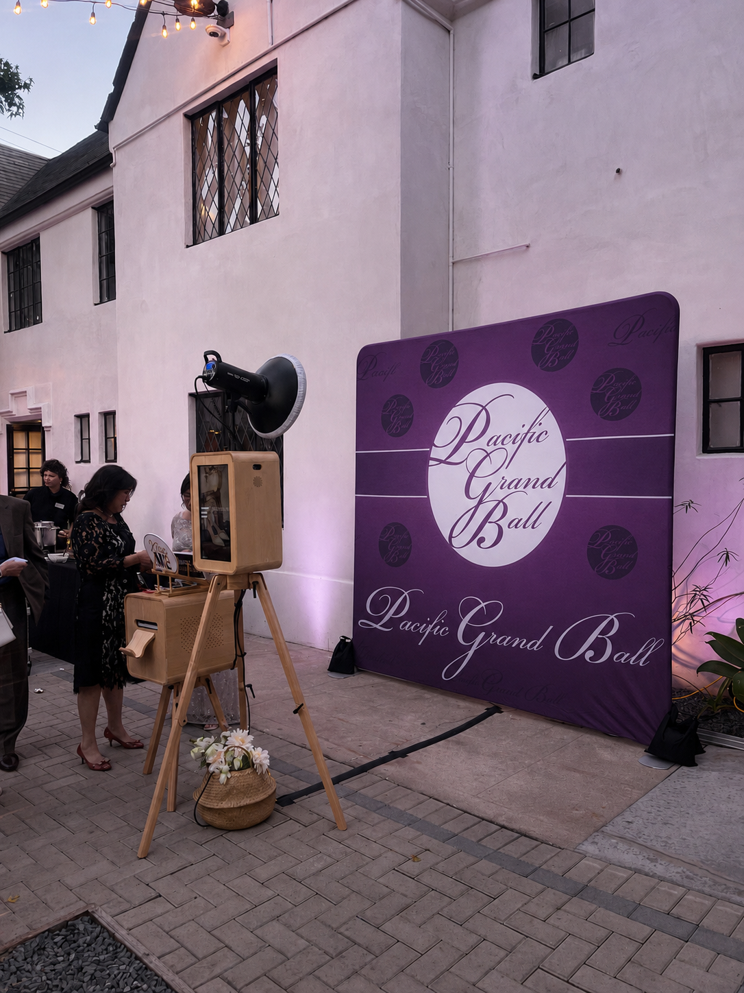 People arriving at a Pacific Grand Ball event outdoors, with a large purple backdrop and string lights overhead.