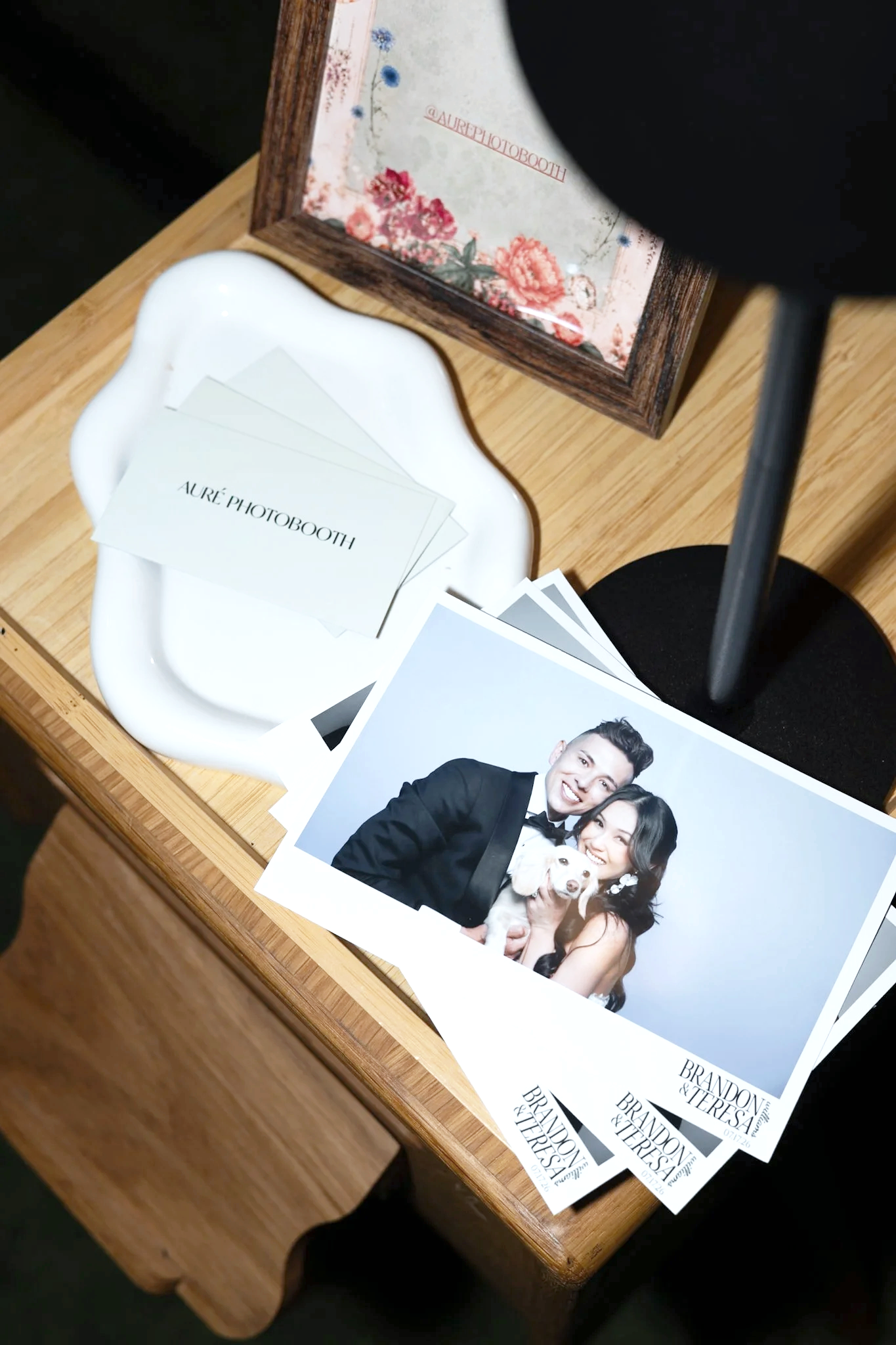 A wooden table holding a stack of wedding photo prints, a white ceramic tray with printed cards that say 'AURÉ PHOTOBOOTH,' and a framed floral artwork that reads '@AURÉPHOTObooth.'