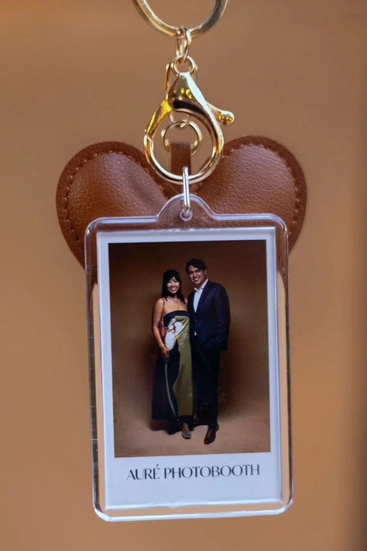 Photo booth photo featuring a smiling woman in a black and yellow dress and a smiling man in a dark suit, standing together against a brown background. The photo is attached to a keychain with a gold clasp, on a heart-shaped leather background, labeled 'AURÉ PHOTOBOOTH.'