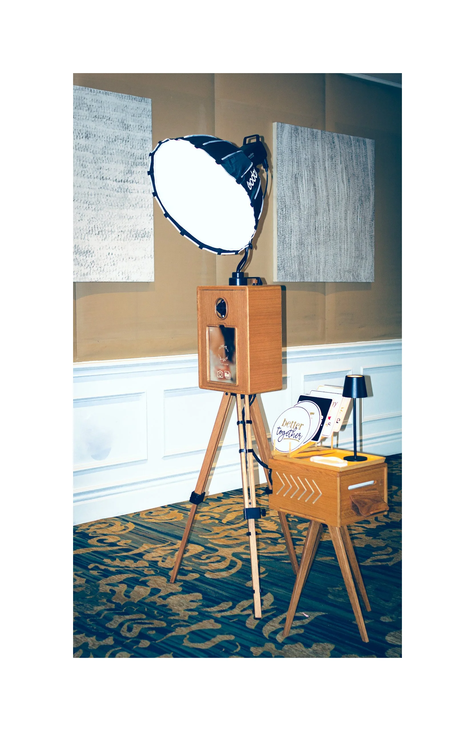 Photo of a photography setup in a room with beige walls. There is a large reflective umbrella attached to a stand, a wooden tripod, a small wooden table with a lamp, framed artwork on the wall, and a patterned carpet.