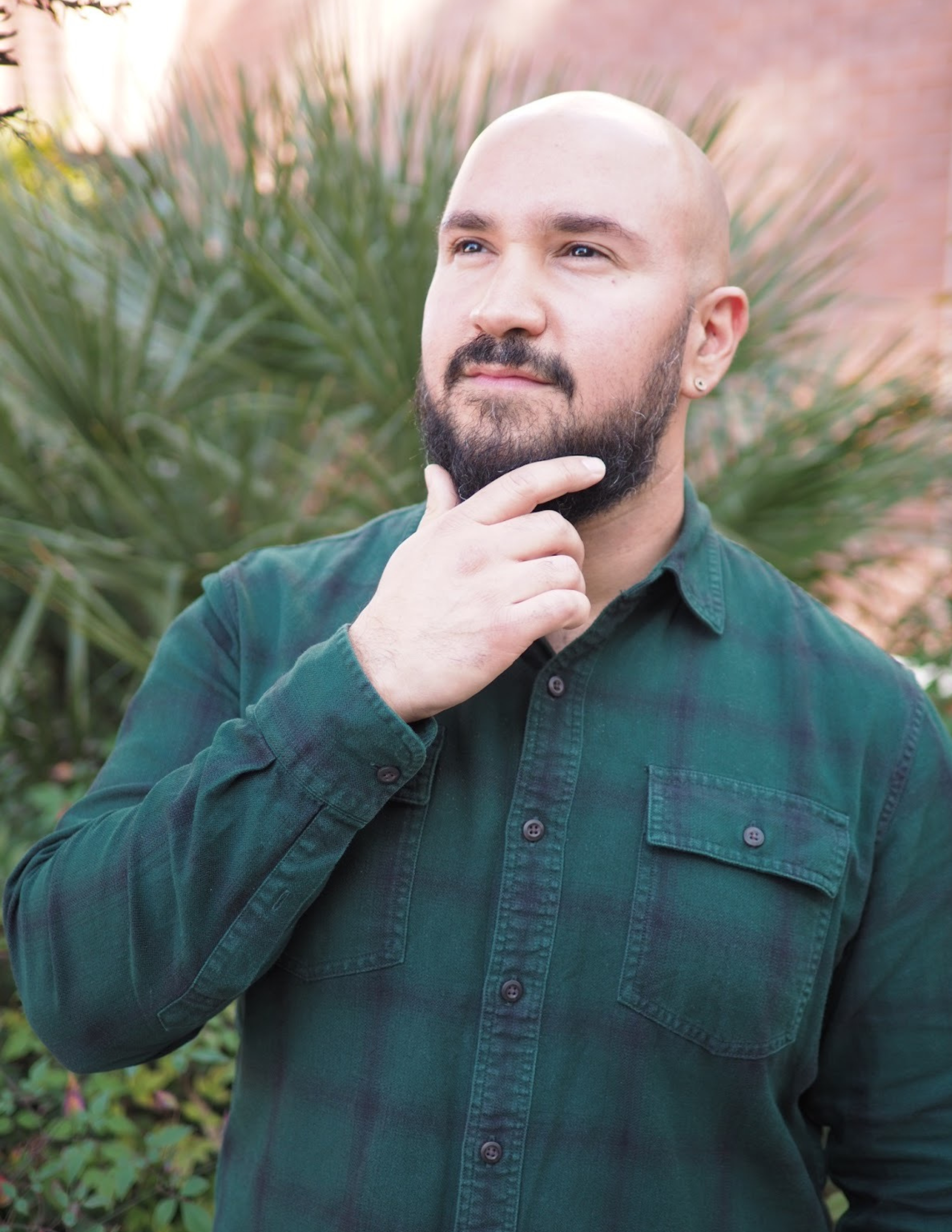 A man with a beard and shaved head, wearing a green shirt, standing outside with greenery and a brick wall in the background, looking thought-provoking with his hand on his chin.