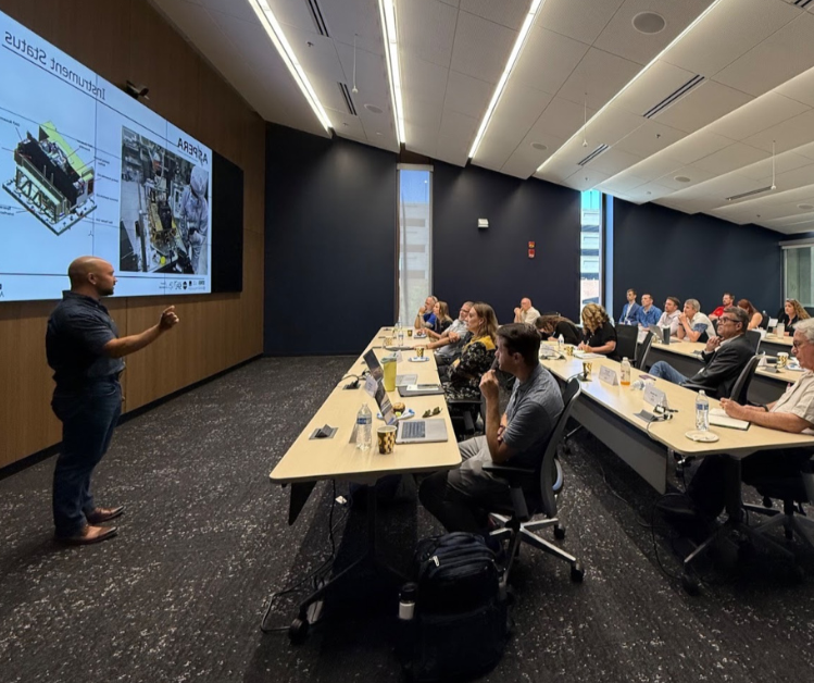 A presenter standing and pointing at a large screen displaying technical diagrams, with an audience of diverse adults seated at conference tables in a modern room.