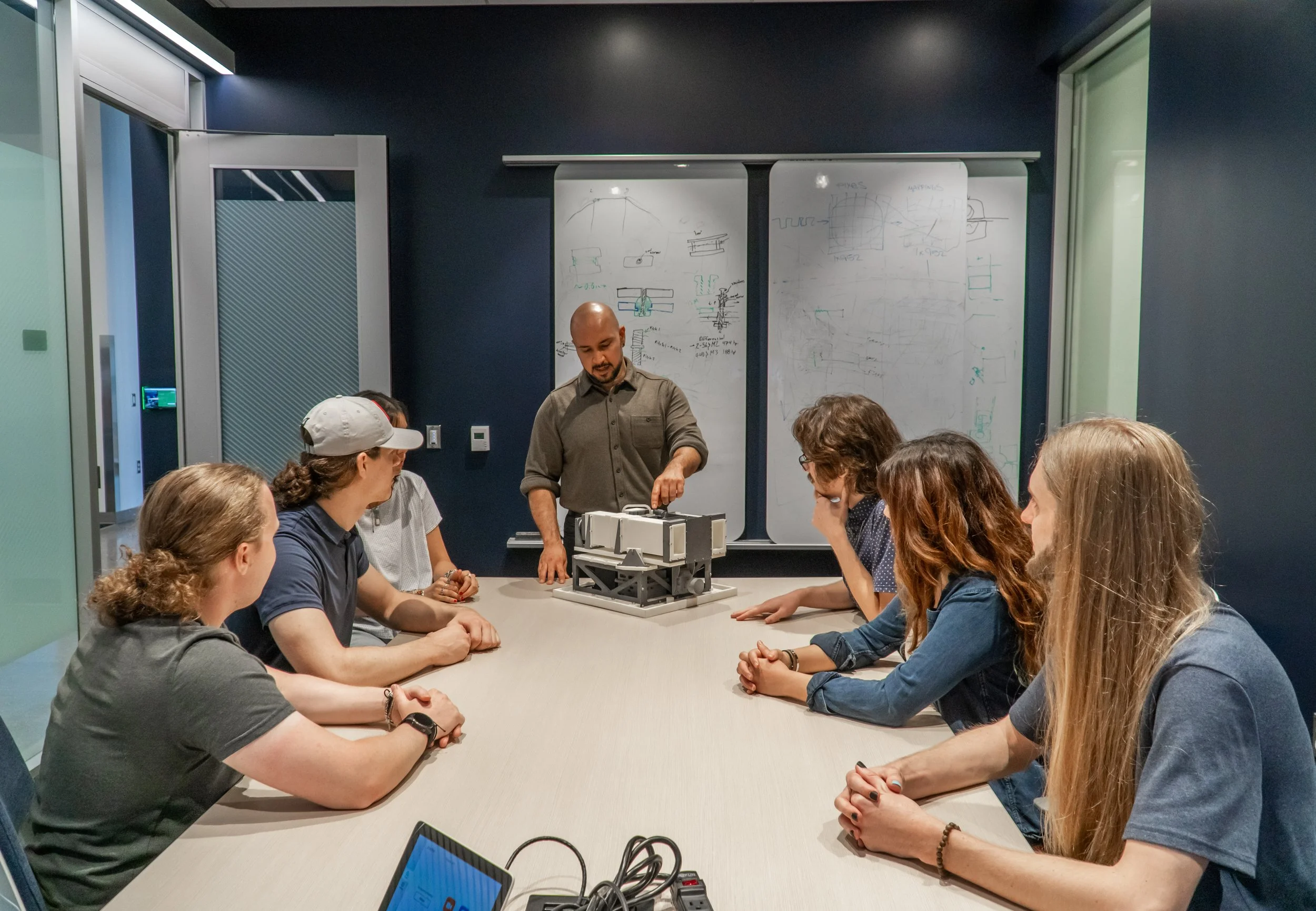 A group of seven students sit around a conference table in a classroom, listening to an instructor who is pointing to a large, complex mechanical or electronic device on the table. The background features whiteboards with technical drawings and notes.