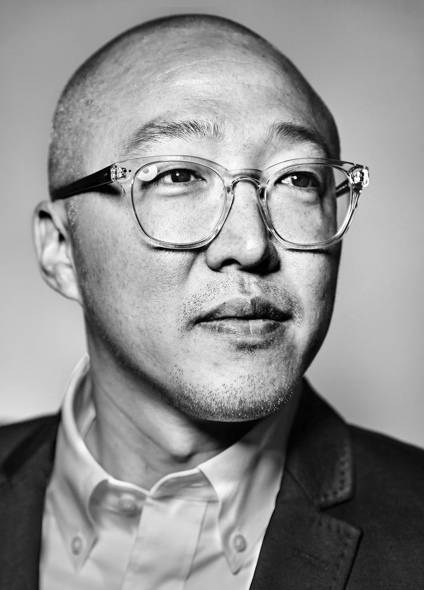 Black and white portrait of a man with a shaved head, wearing large clear glasses, a white collared shirt, and a dark blazer.