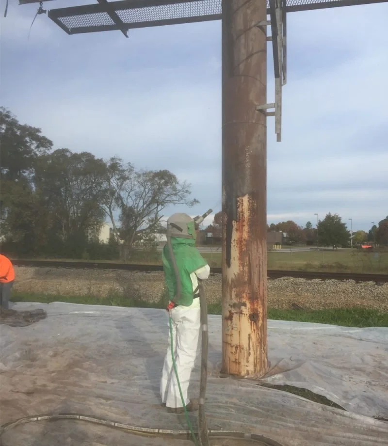 Worker in protective gear sandblasting a rusted utility pole outdoors with a cloudy sky and trees in the background.