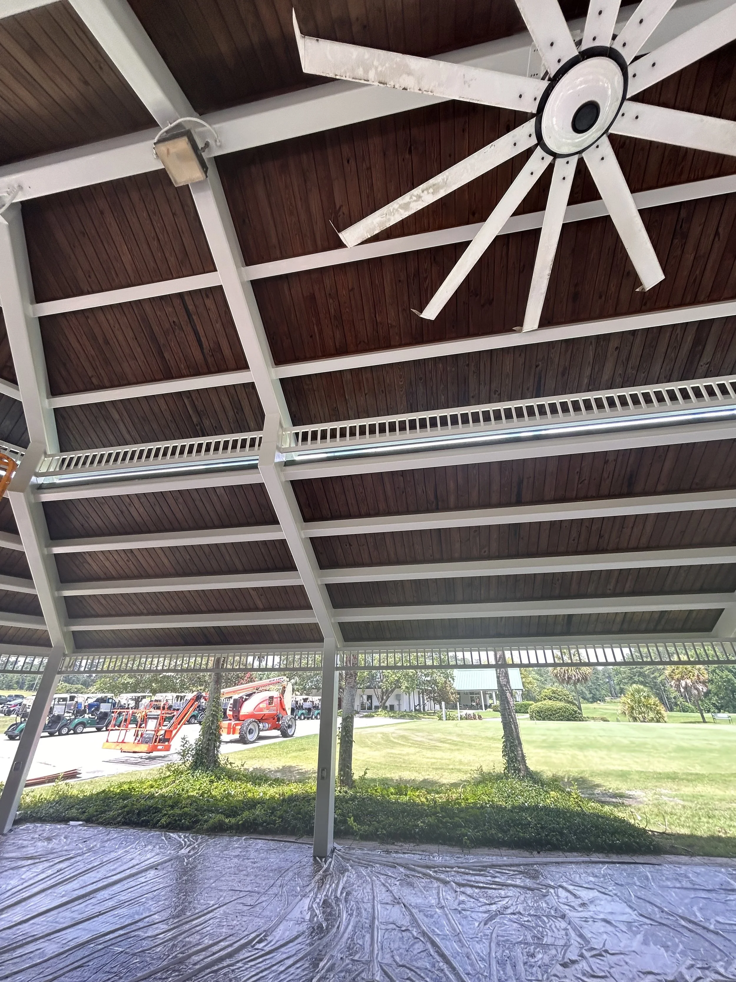 View of a covered outdoor area with a ceiling fan, construction equipment, and a grassy landscape with trees and buildings in the background.