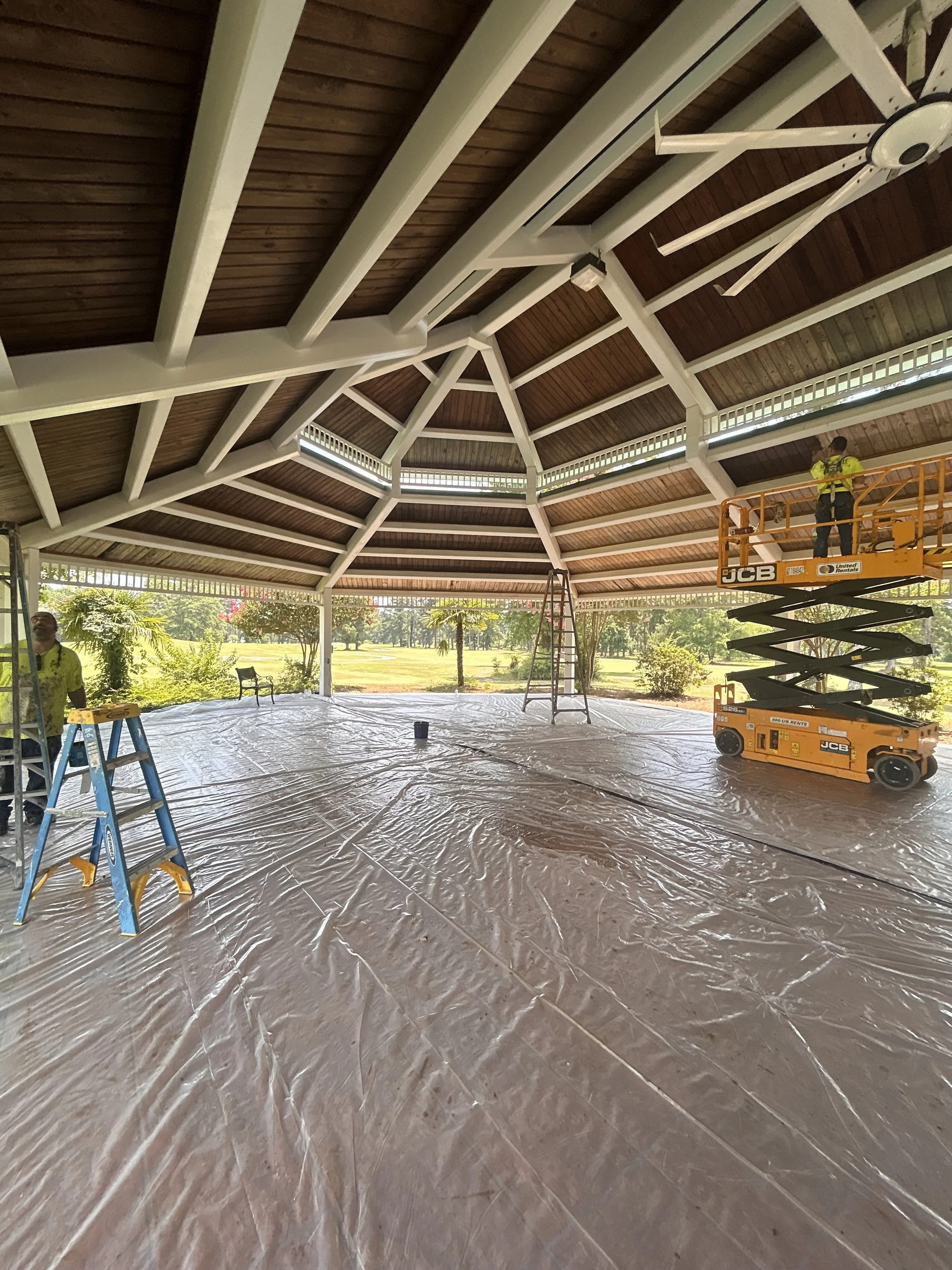 Construction workers installing a wooden ceiling in an open pavilion with scaffolding and ladders, surrounded by trees and a grassy area.