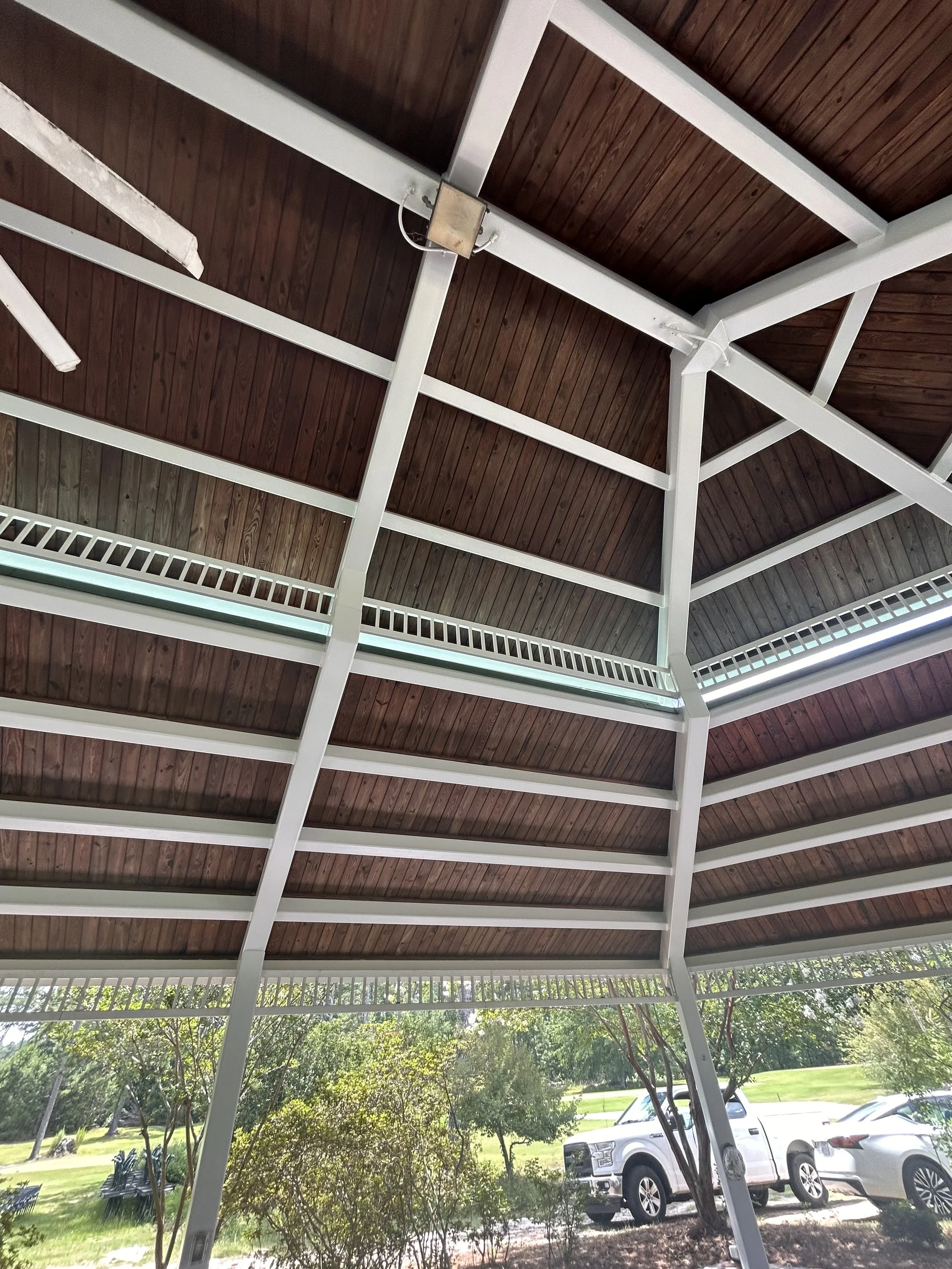 View of a parking lot with several cars, surrounded by trees and greenery, under a wooden and white metal shelter roof.