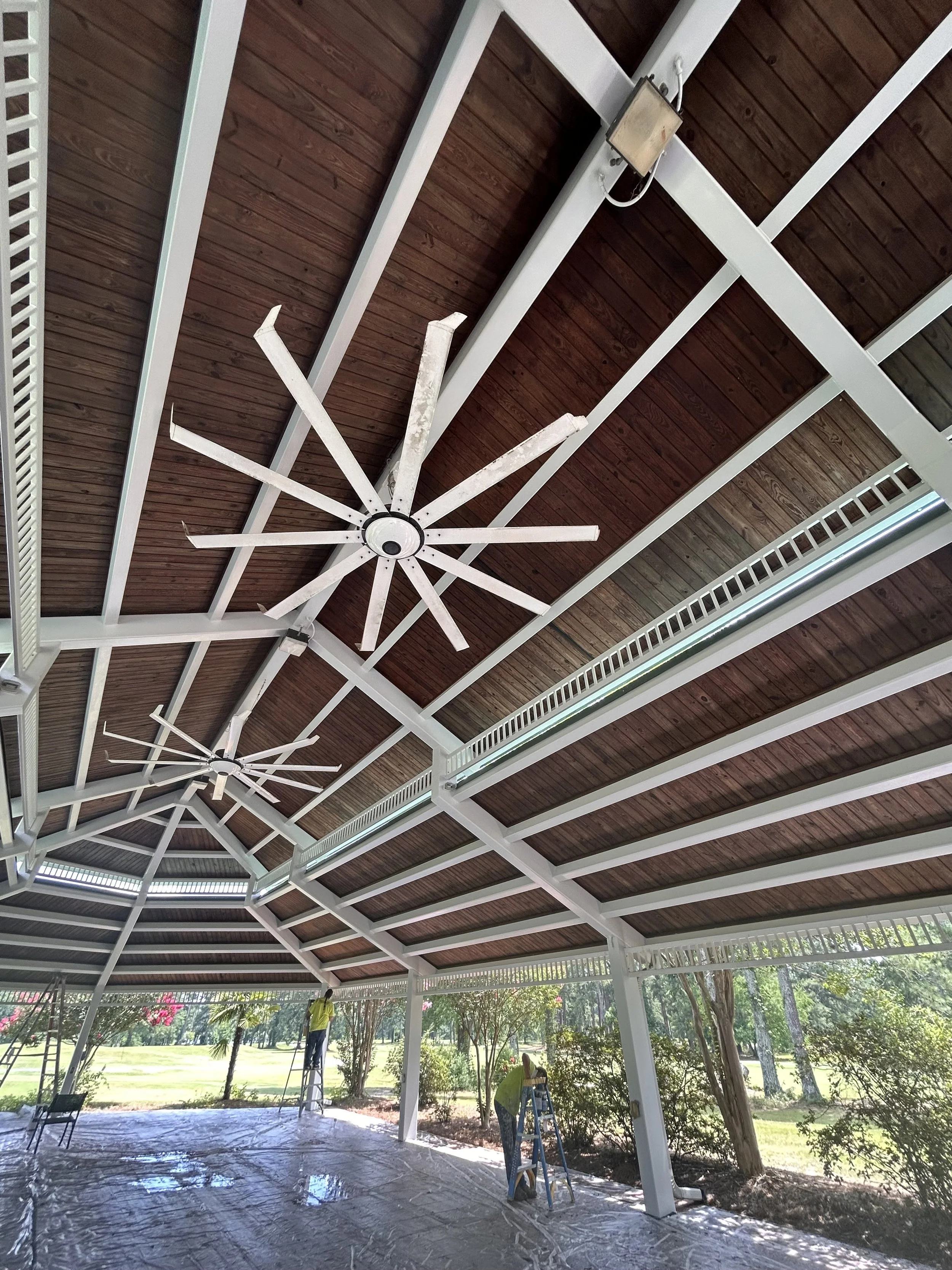 A covered outdoor patio with a ceiling fan under construction, showing workers, a ladder, and trees in the background.