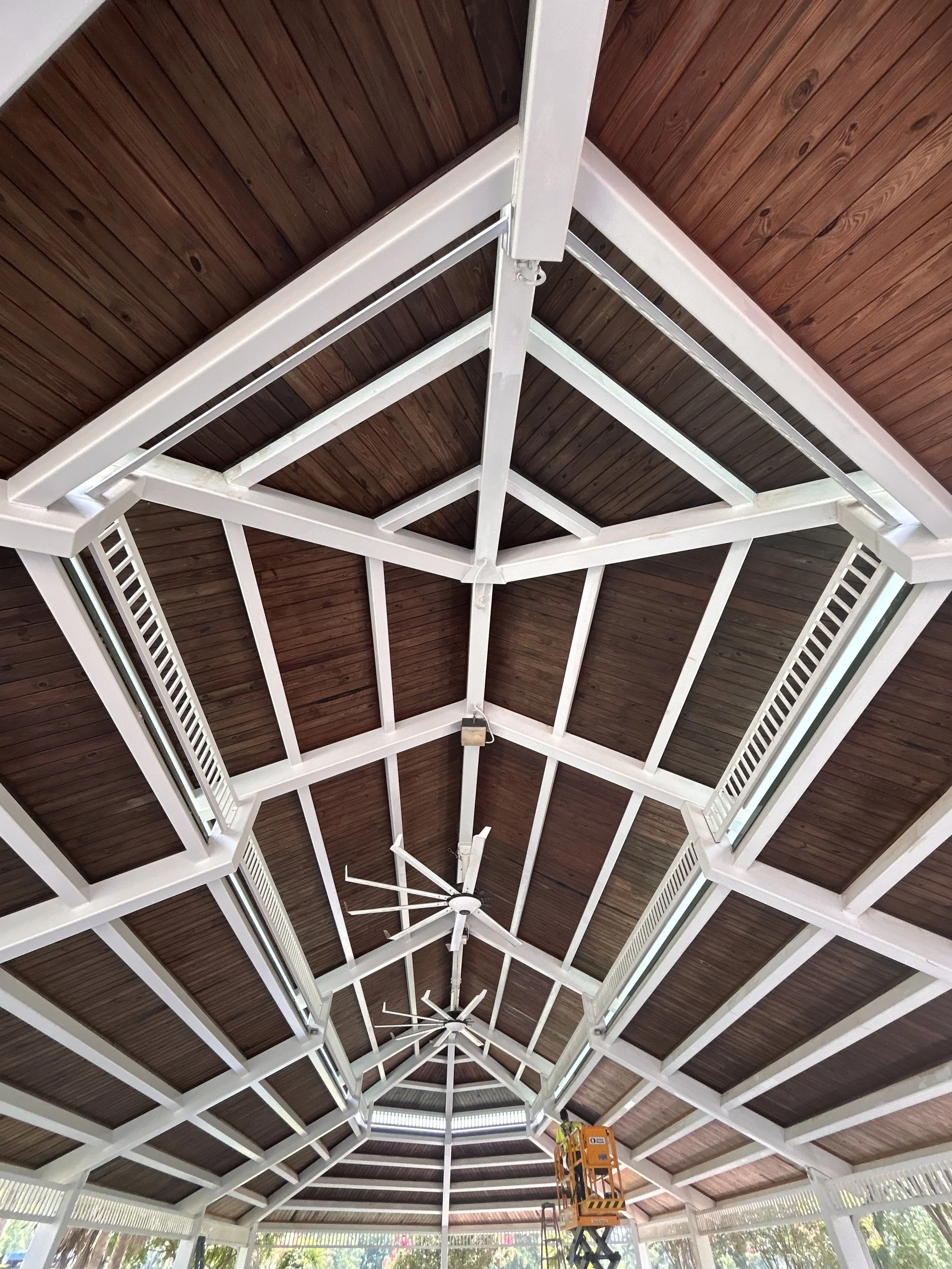 Looking up at a gazebo ceiling with dark wooden planks and white painted beams, including a hanging ceiling fan and a worker in a cherry picker at the bottom.