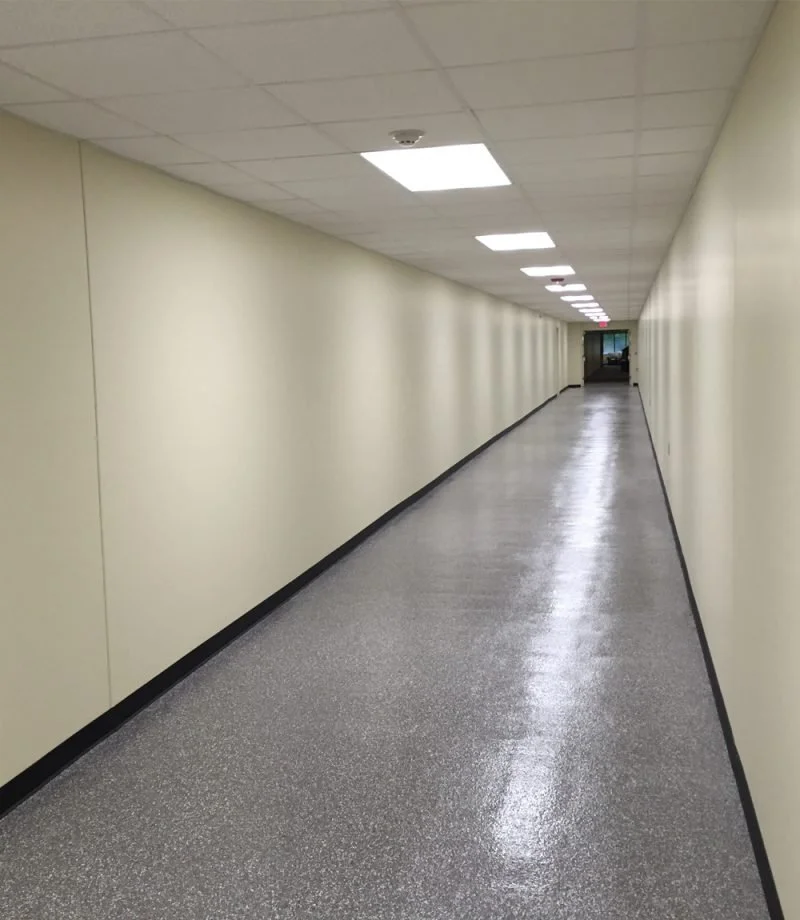 Long indoor hallway with beige walls, gray speckled floor, and fluorescent ceiling lights extending to a closed door at the end.