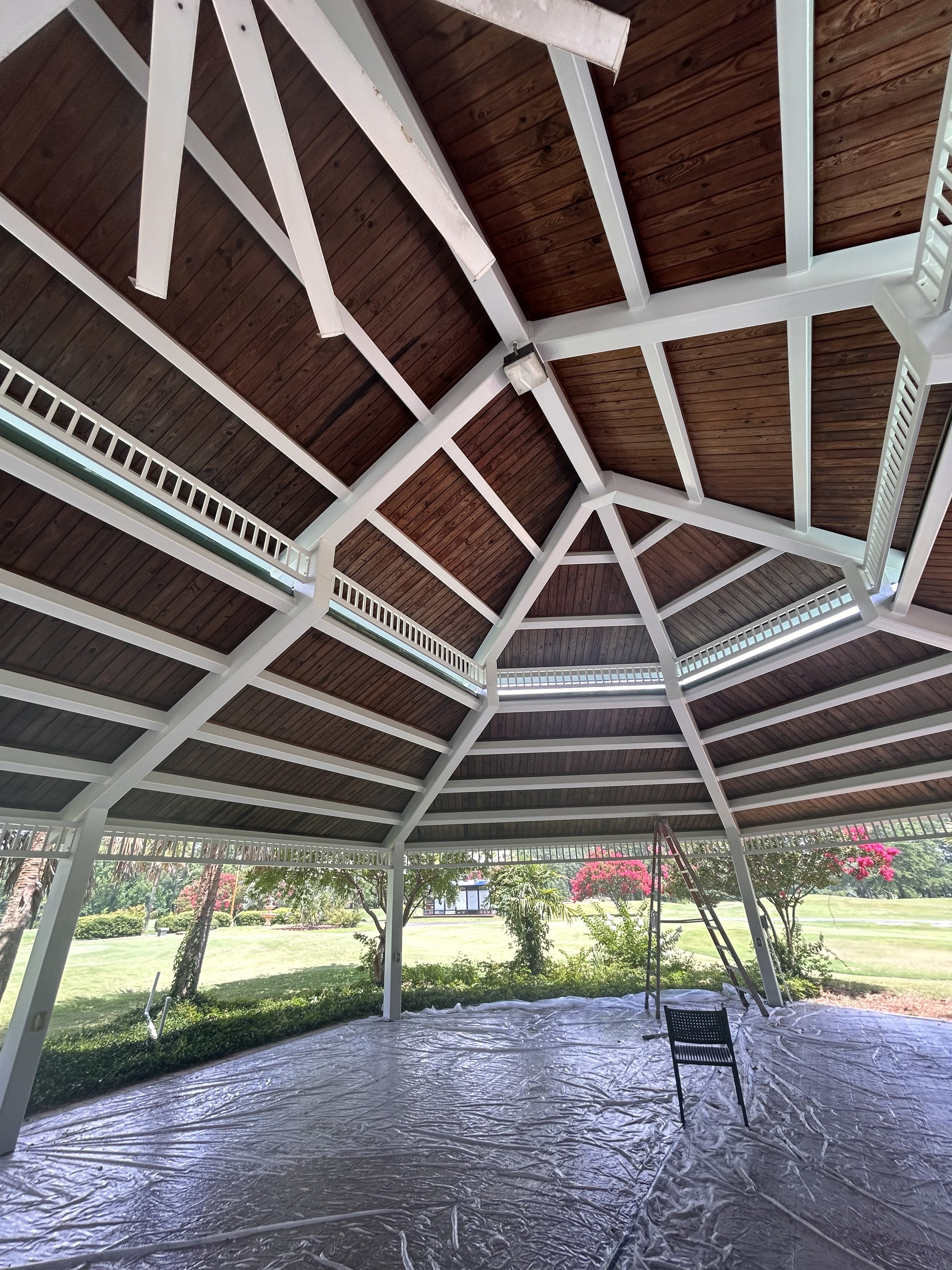 The ceiling of a gazebo with a wooden interior, white framing, and a lantern light fixture, surrounded by landscaping with trees and flowering bushes, and covered with a plastic tarp.