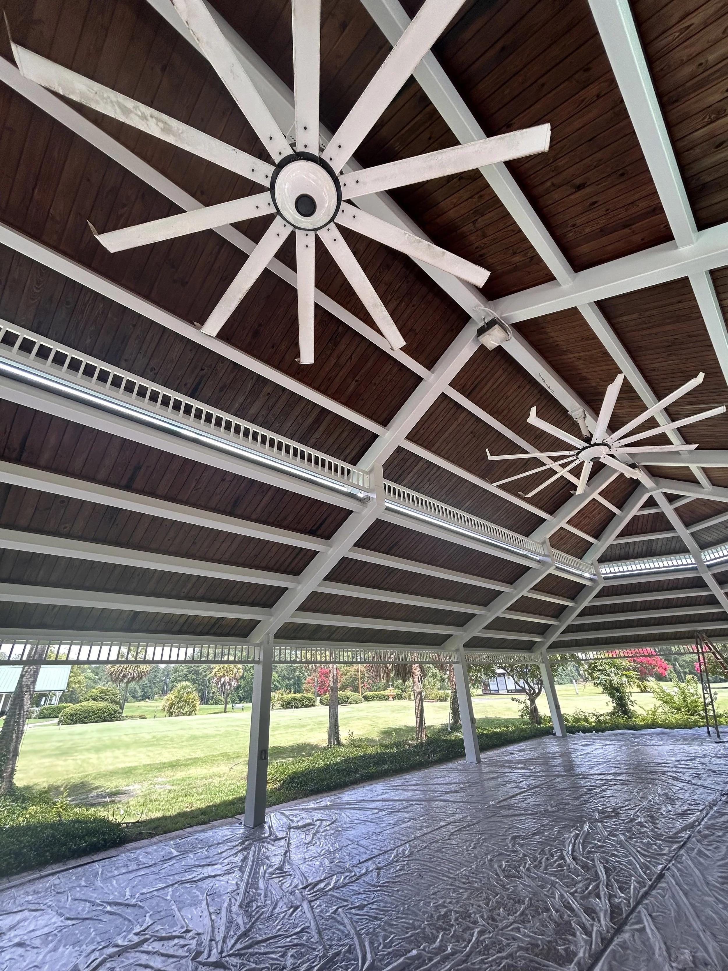 The ceiling of a pavilion with two white ceiling fans and a wooden roof. Outside, there is a grassy area with trees and pink flowers.
