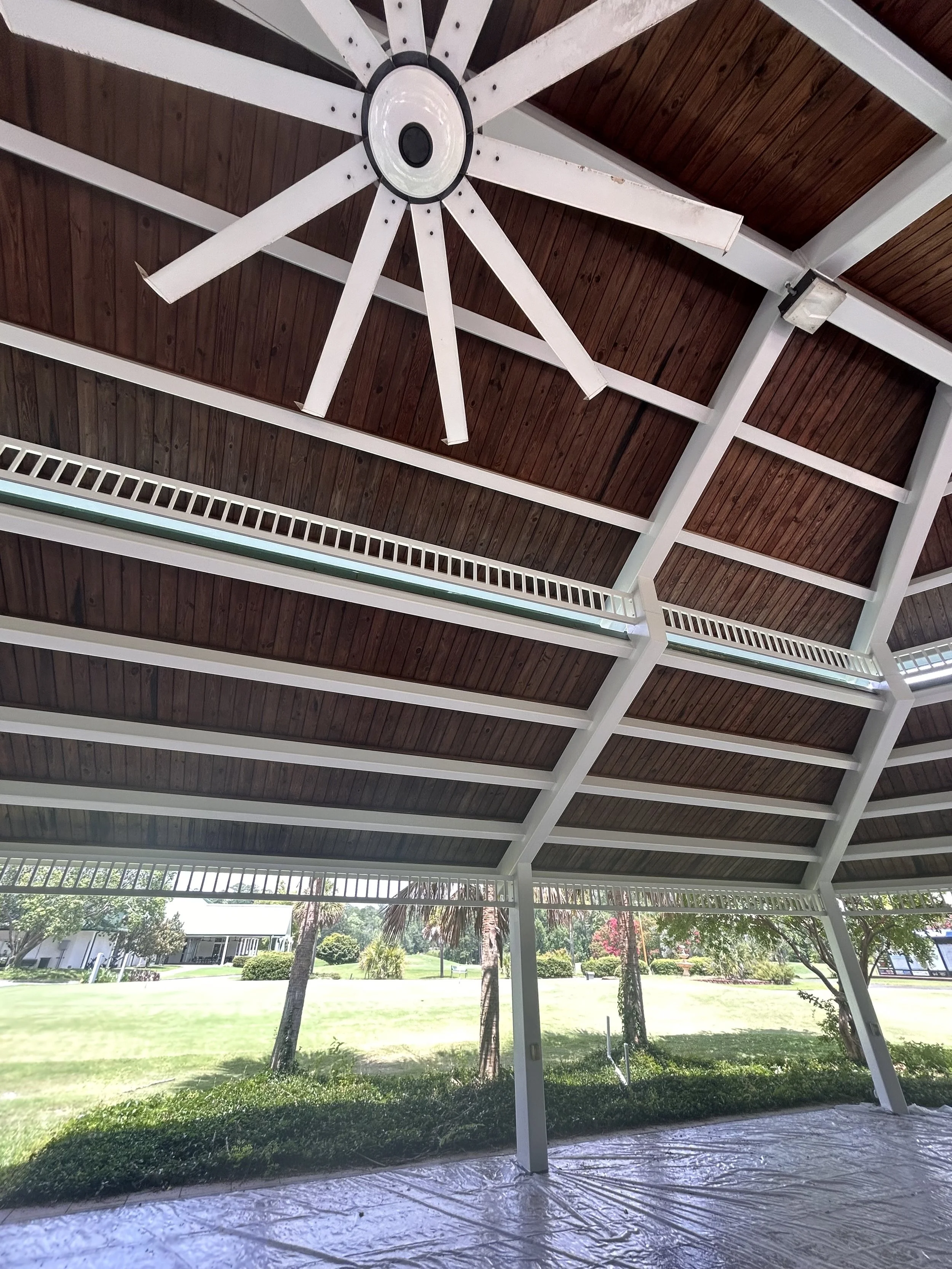 Ceiling fan hanging from a wooden ceiling of a covered porch area with an outdoor scene including trees and a lawn in the background.