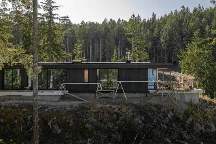 Dark clad house with a long run of glazing, sawhorses on a concrete terrace, forest behind
