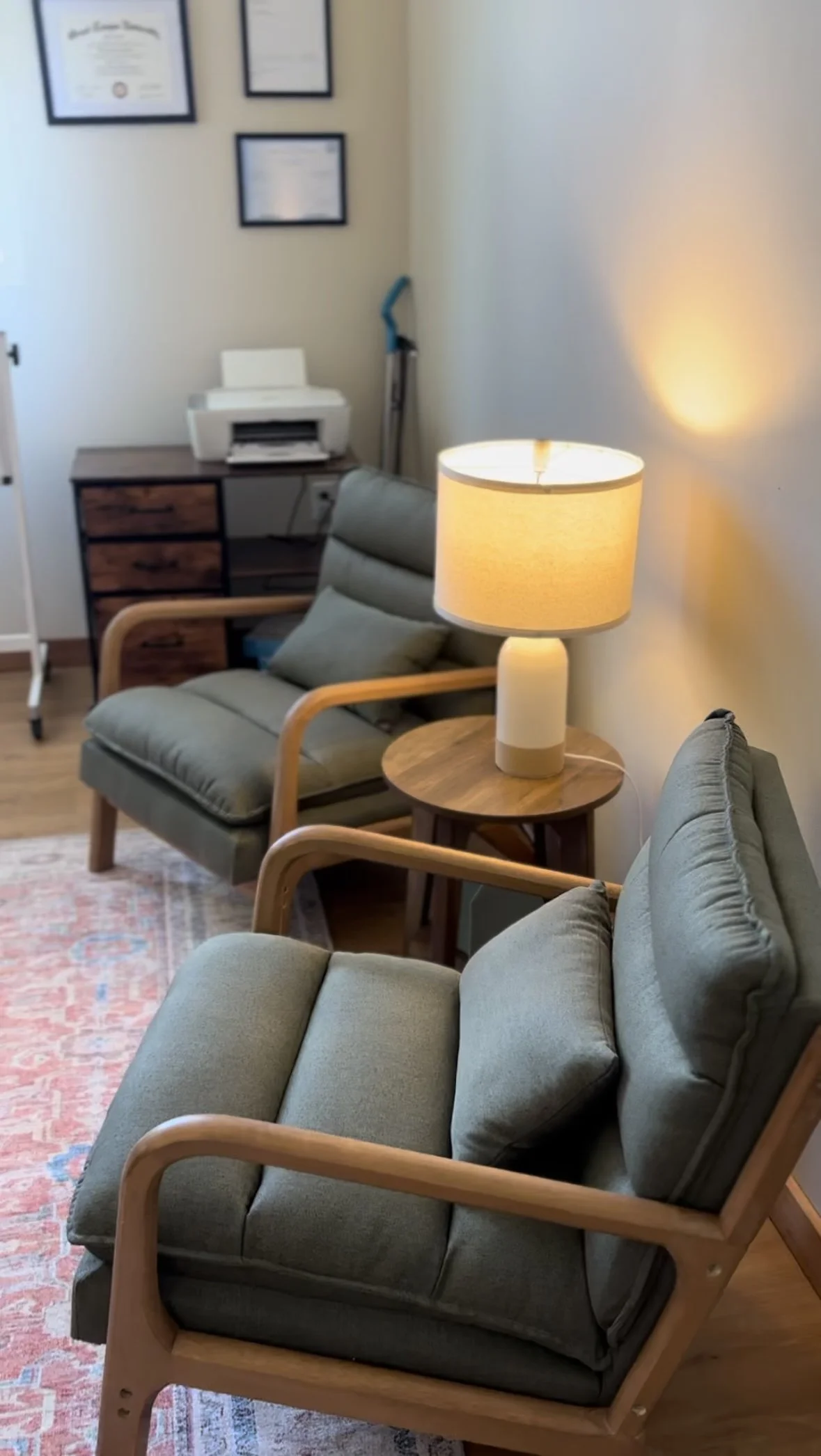Living room with two green armchairs, wooden armrests, a small round wooden side table with a glowing table lamp, a patterned rug, a desk with framed certificates on the wall behind, a printer and a vacuum cleaner in the corner.