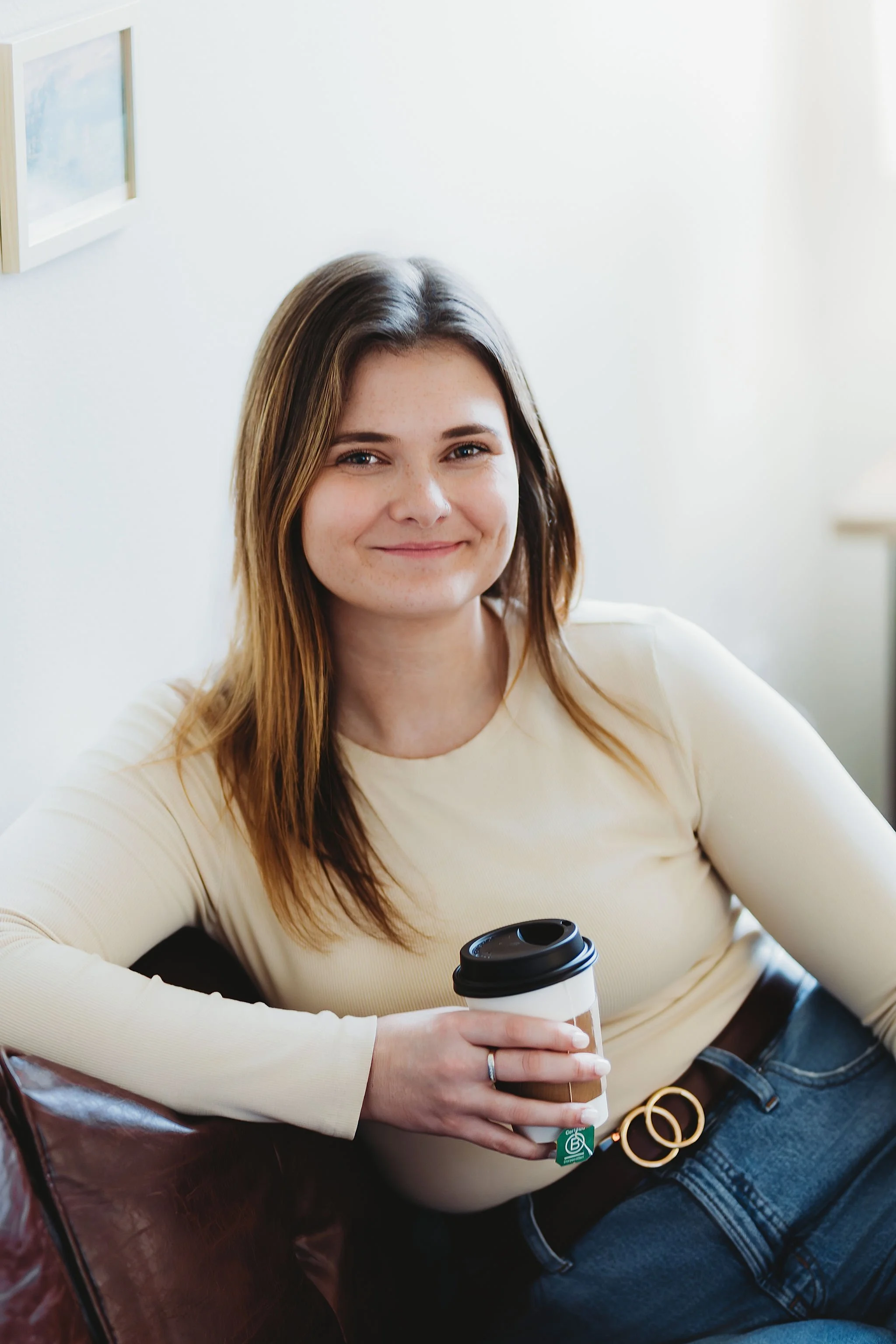 A young woman with brown hair and freckles smiling, sitting with a takeaway coffee. She is wearing a cream-colored top, dark blue jeans, and a gold ring, in a bright indoor setting.