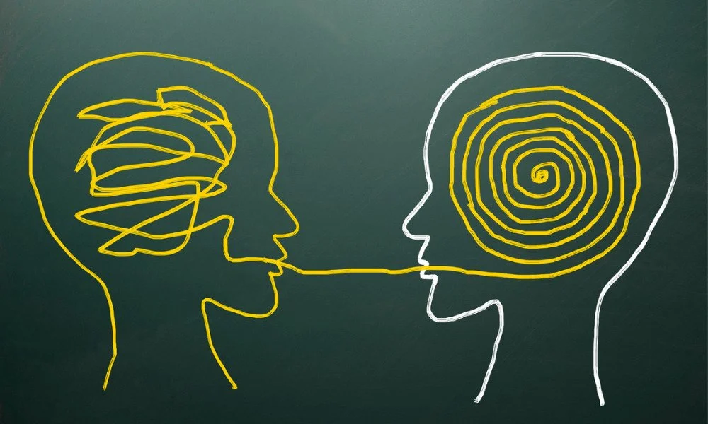Illustration of two human profiles facing each other with their brains depicted as abstract, colorful head diagrams; one in yellow with a scribbled pattern and the other in white with a spiral pattern, connected by a straight line suggesting communication or thought transfer.