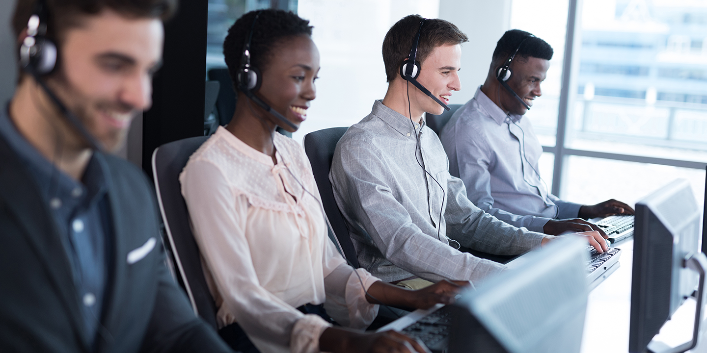 Four customer service representatives in an office, wearing headsets, working at their computers with large windows in the background.