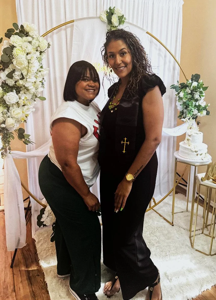 Two women posing for a photo in front of a gold arch decorated with flowers