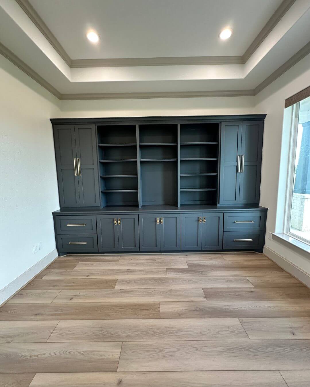 A modern built-in dark gray bookshelf with closed cabinets at the bottom, open shelves in the middle, situated in a room with light-colored wall and wood plank flooring. A window on the right lets in natural light.