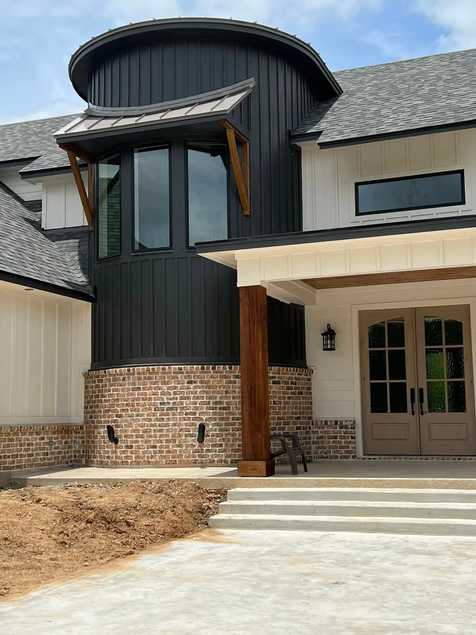 Modern house with black metal siding, brick foundation, and a porch with steps, featuring a large bay window with a small roof and a glass double door.