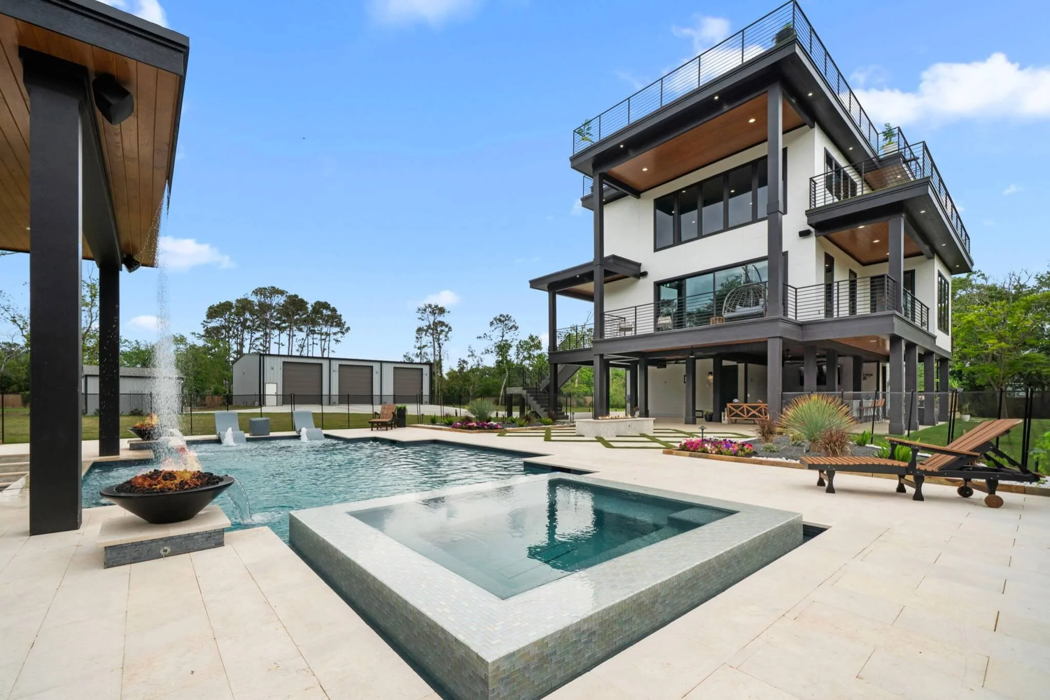 Modern multi-story house with large balconies overlooking a backyard with a swimming pool, fire pit, and outdoor seating area, surrounded by trees and a clear blue sky.