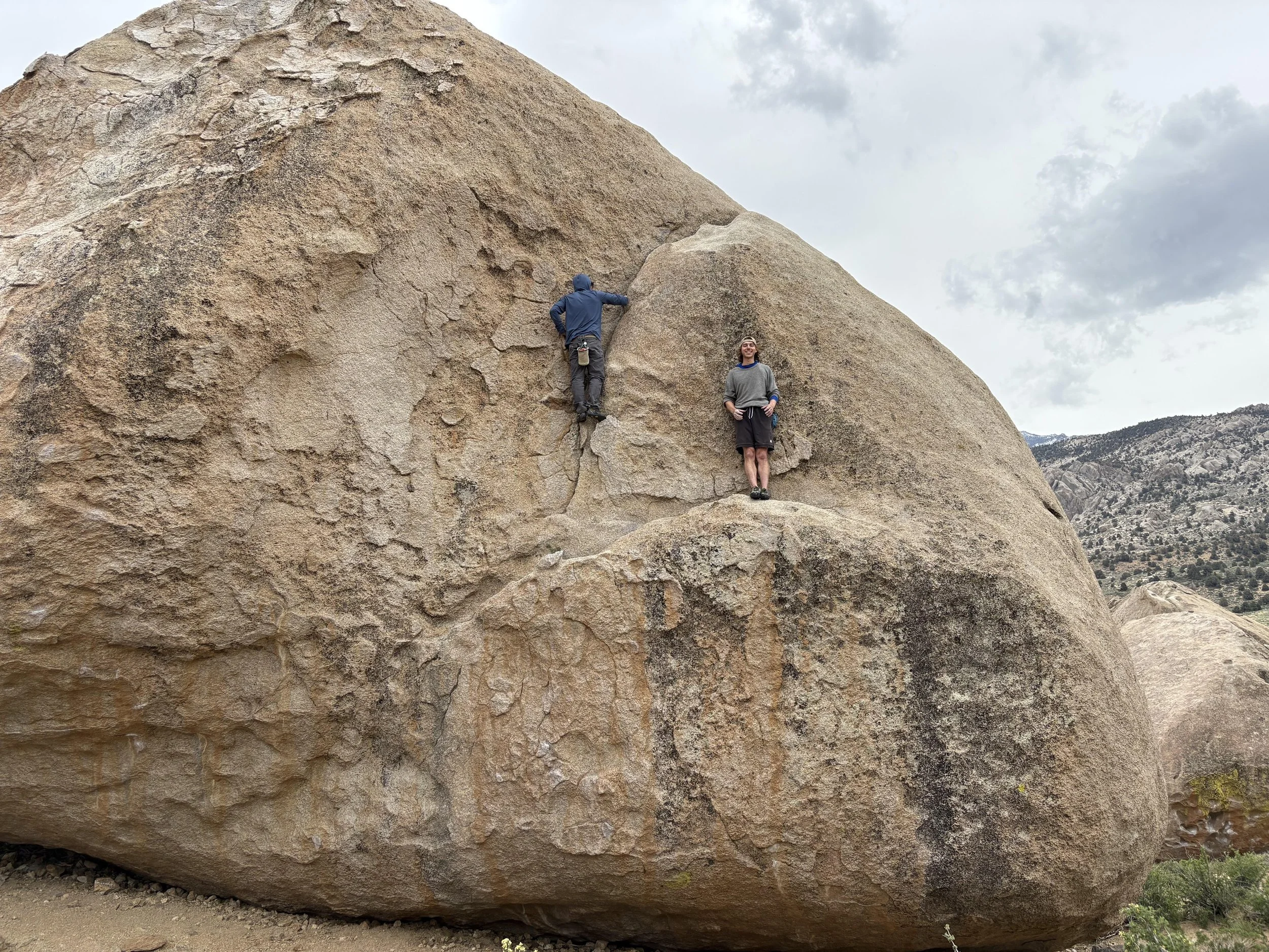 Two climbers on a large boulder, one climbing and one standing on the rock outdoors with a mountainous background and cloudy sky.