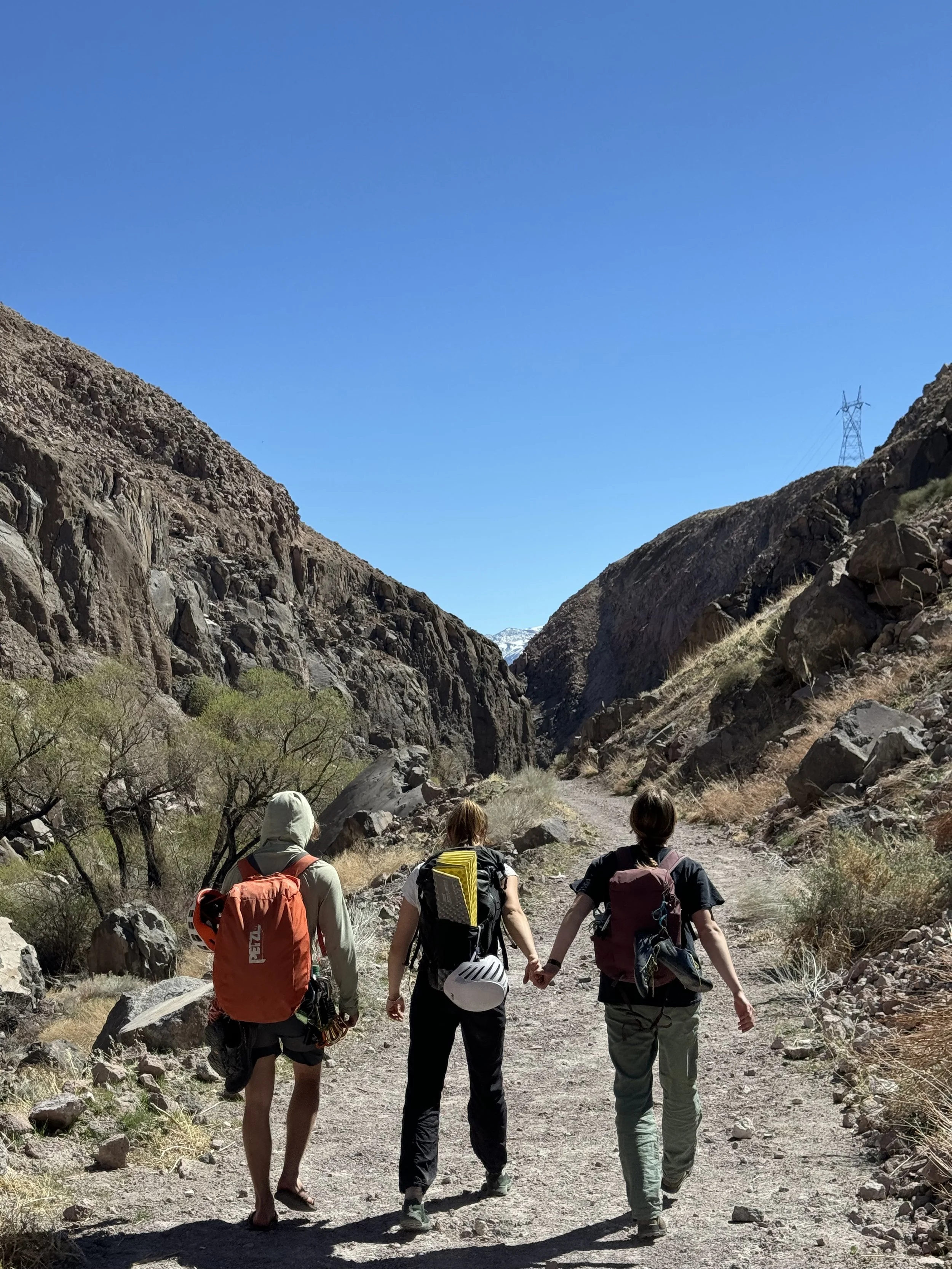 Three hikers walking through a rocky canyon on a clear sunny day, two women holding hands, with rugged hills and sparse vegetation around.