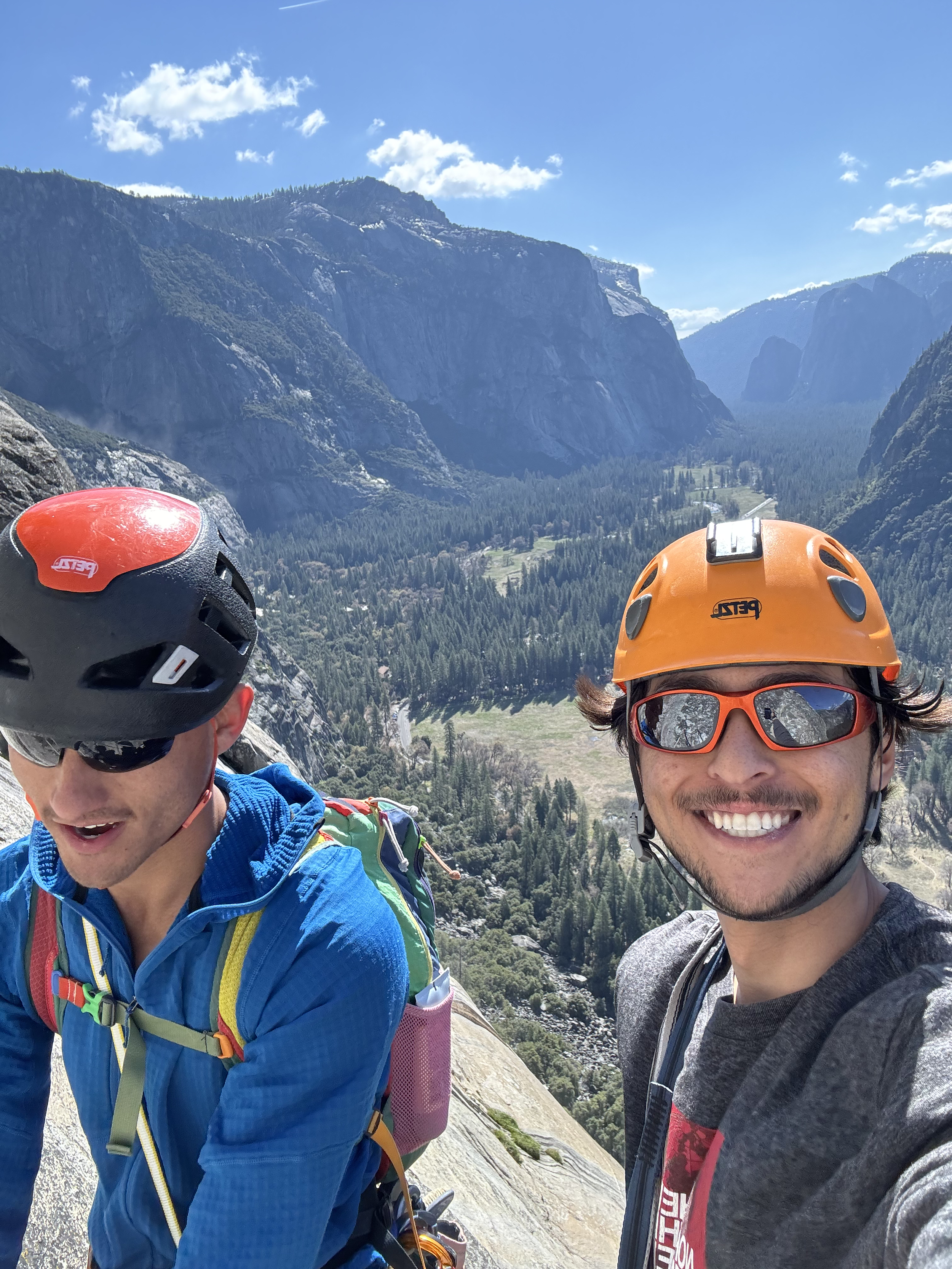 Two men wearing helmets and sunglasses taking a selfie on a mountain with a lush valley and cliffs in the background.