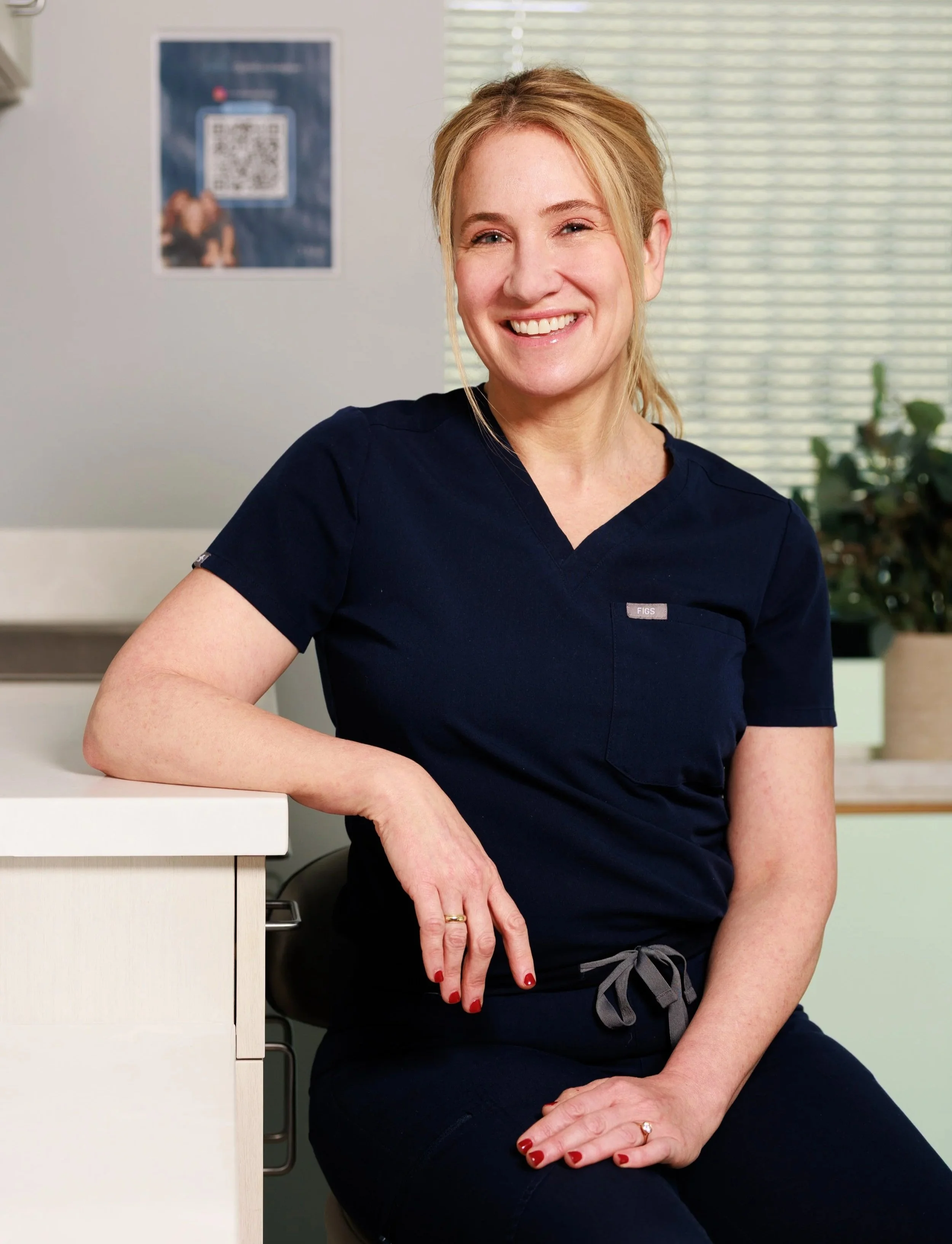 A woman with blonde hair sitting at a table, smiling at the camera, wearing a white top and accessories on her wrists and finger.