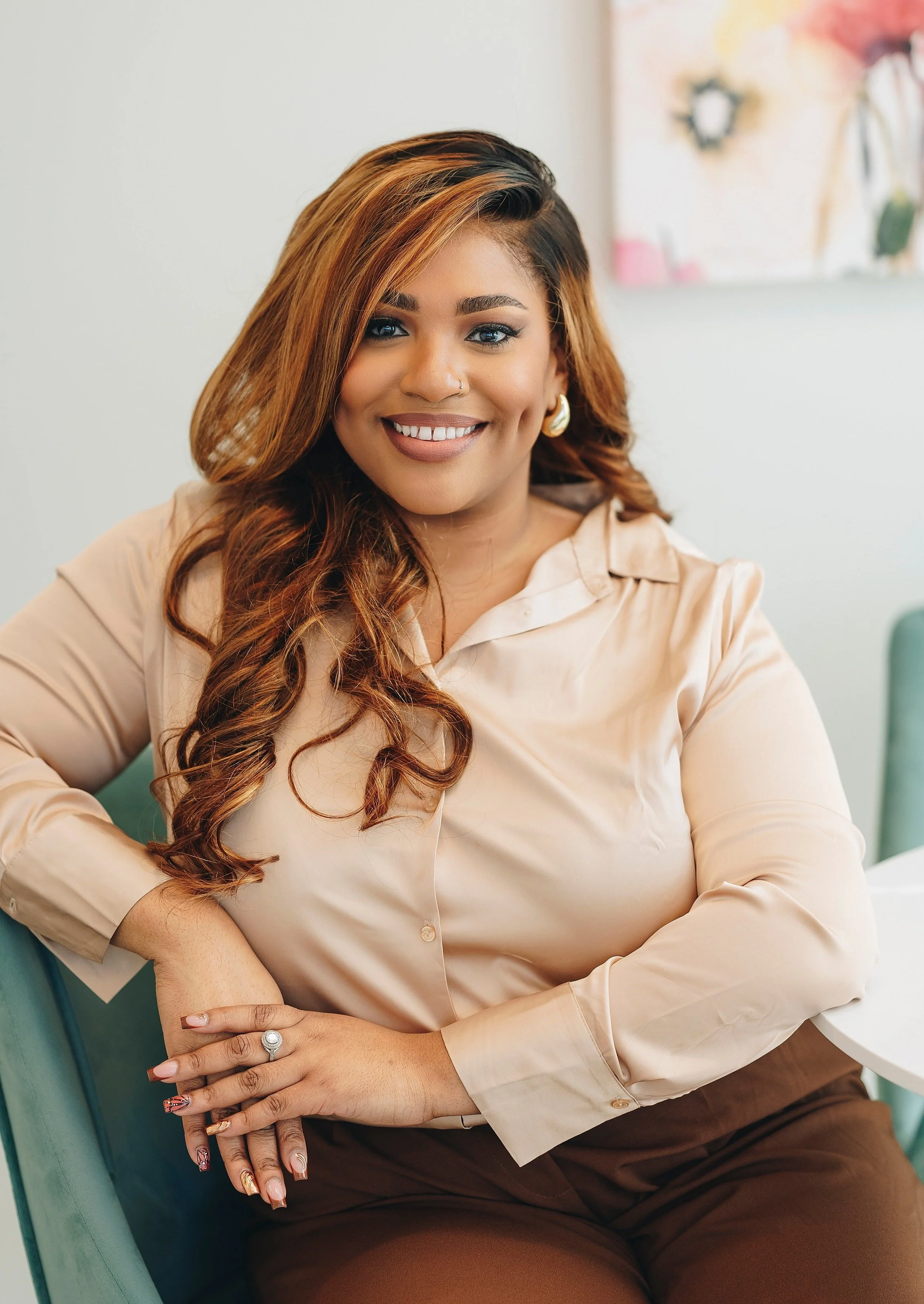 A woman with long, wavy, reddish-brown hair smiling at the camera, wearing a beige blouse and brown pants, sitting at a table in a bright room.