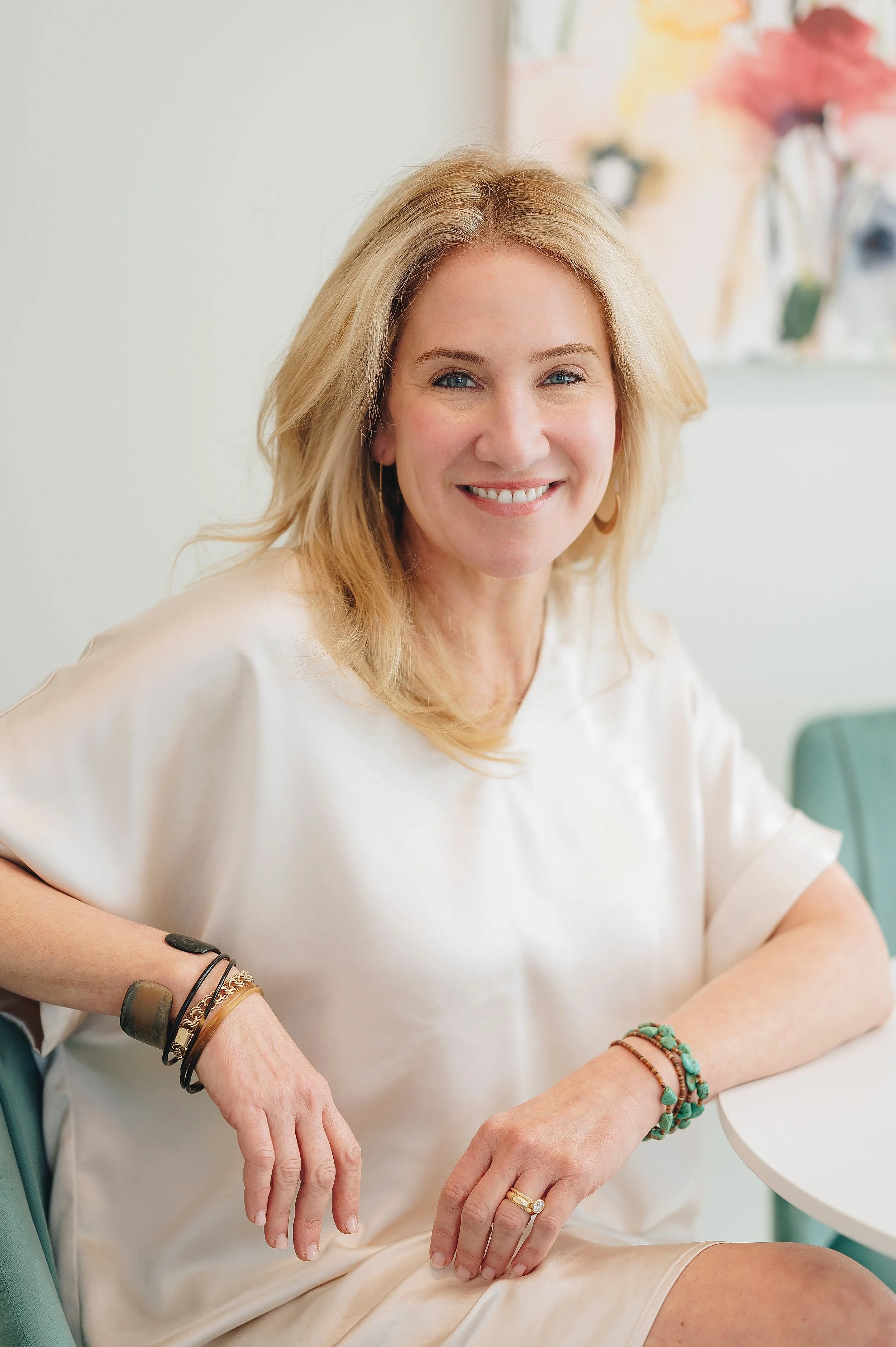 A woman with blonde hair sitting at a table, smiling at the camera, wearing a white top and accessories on her wrists and finger.
