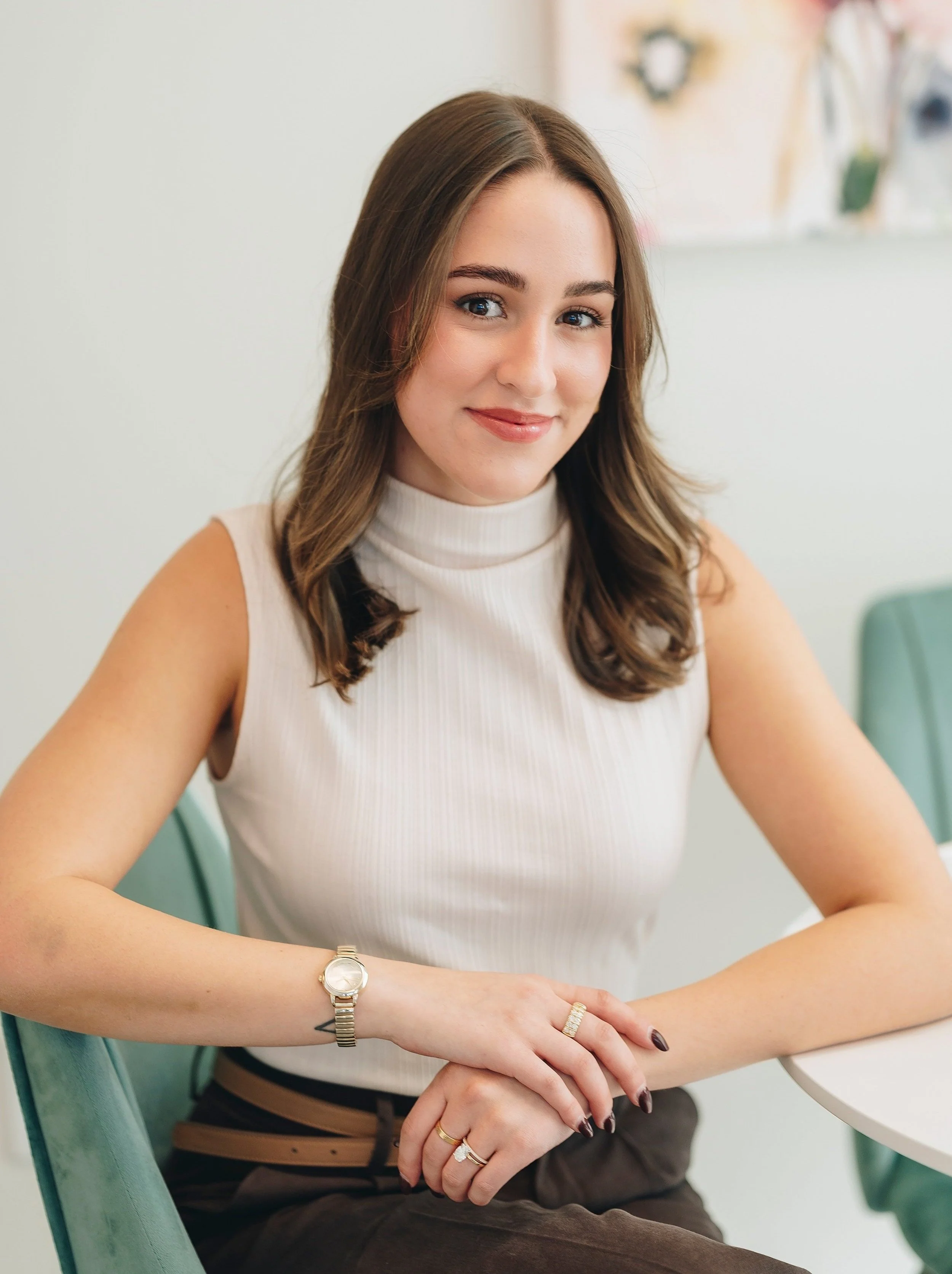 A young woman with shoulder-length brown hair, wearing a sleeveless cream turtleneck top, sitting at a table with her hands resting on the surface, smiling at the camera in a bright room with light-colored walls.