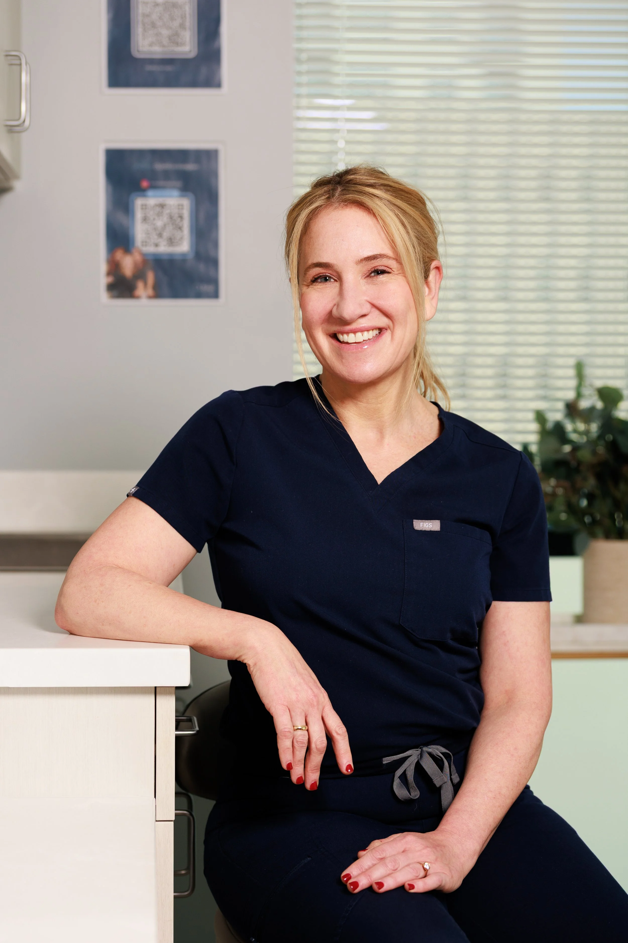 A woman with blonde hair sitting at a table, smiling at the camera, wearing a white top and accessories on her wrists and finger.