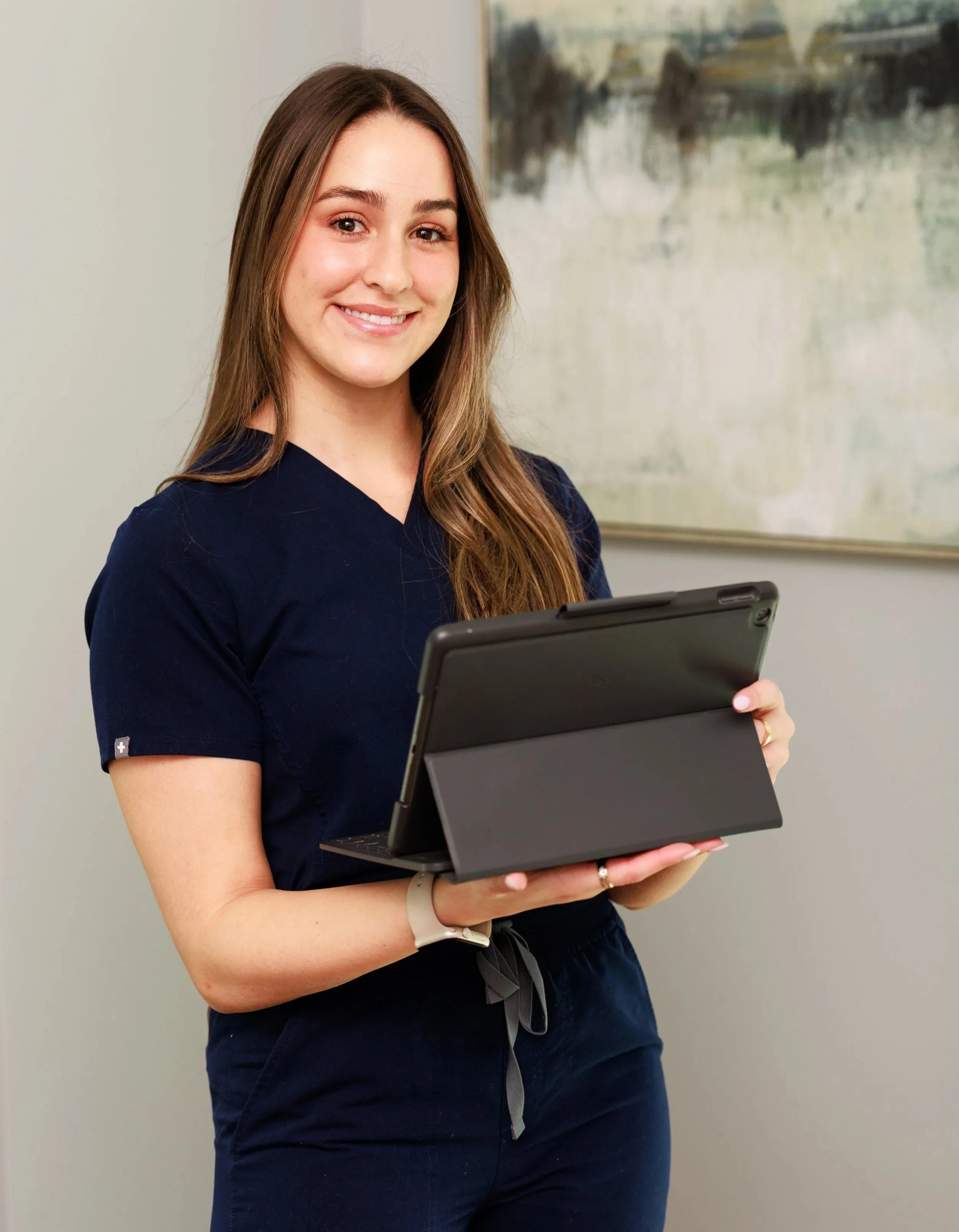 A young woman with shoulder-length brown hair, wearing a sleeveless cream turtleneck top, sitting at a table with her hands resting on the surface, smiling at the camera in a bright room with light-colored walls.