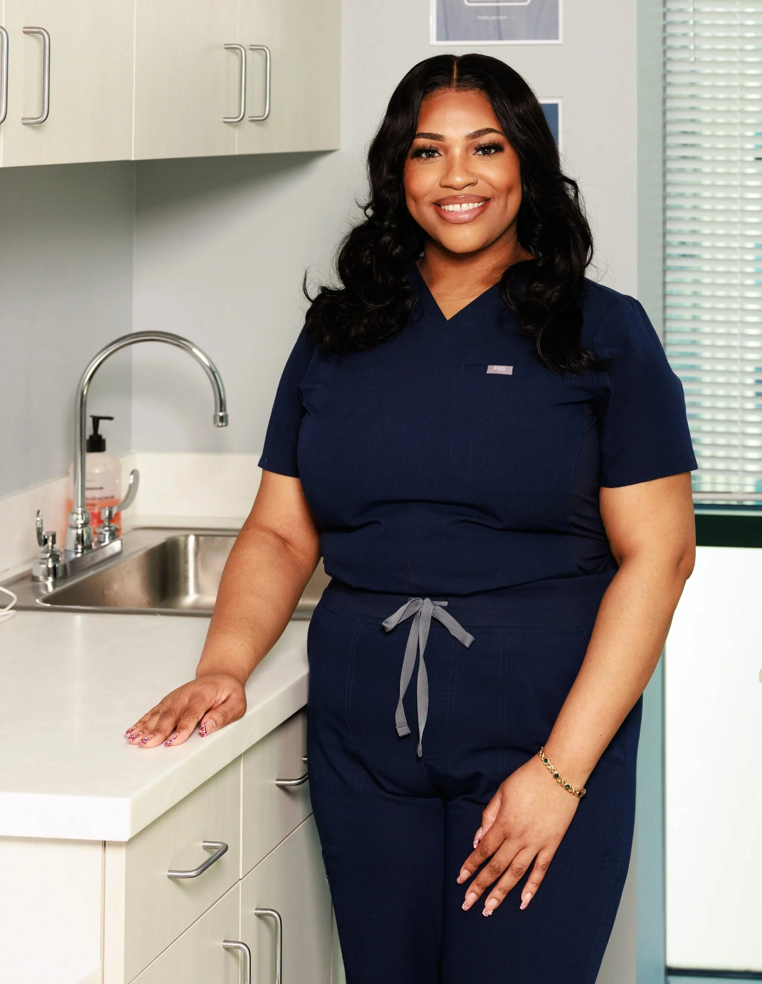 A woman with long, wavy, reddish-brown hair smiling at the camera, wearing a beige blouse and brown pants, sitting at a table in a bright room.