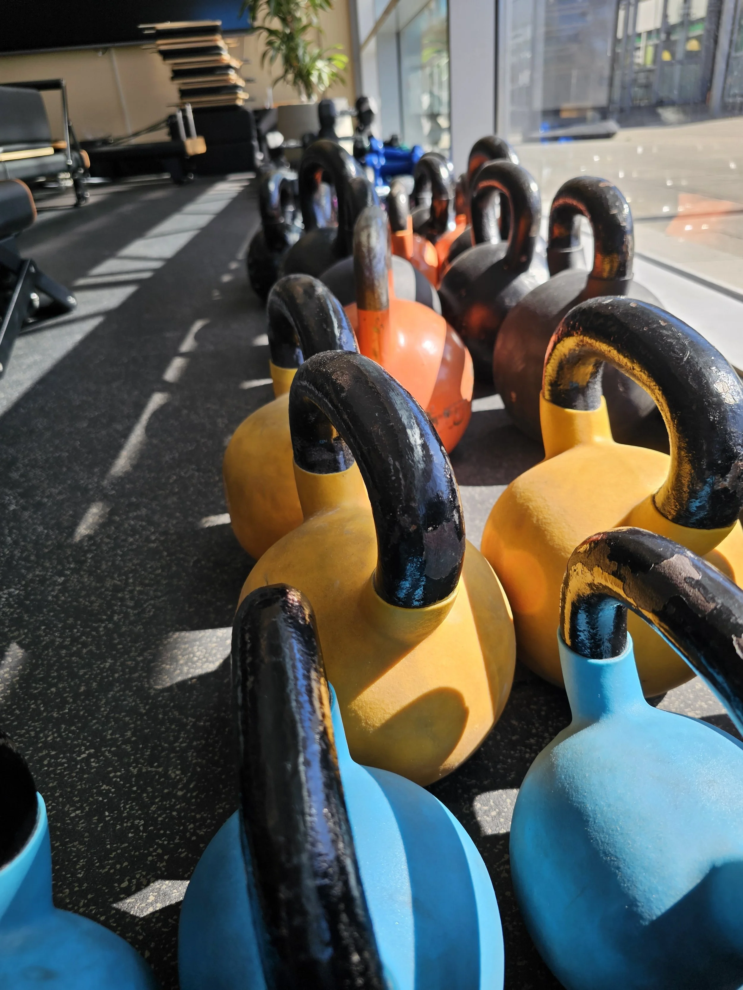 Forskellige farvede kettlebells på en sportscenter gulv, med vindue og udendørsareal i baggrunden.