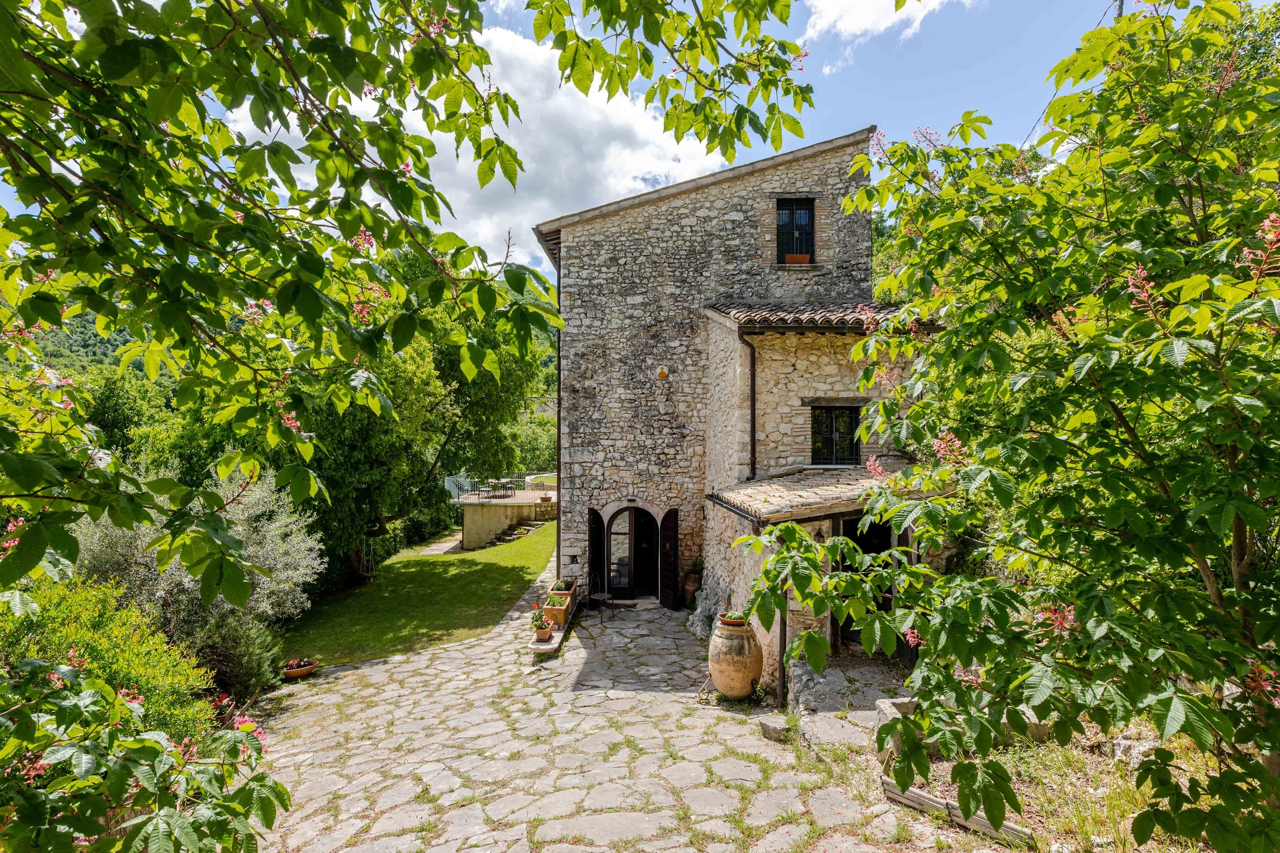 Stone house with arched doorway, surrounded by greenery and trees, with a cobblestone pathway leading up to it, under a partly cloudy sky.