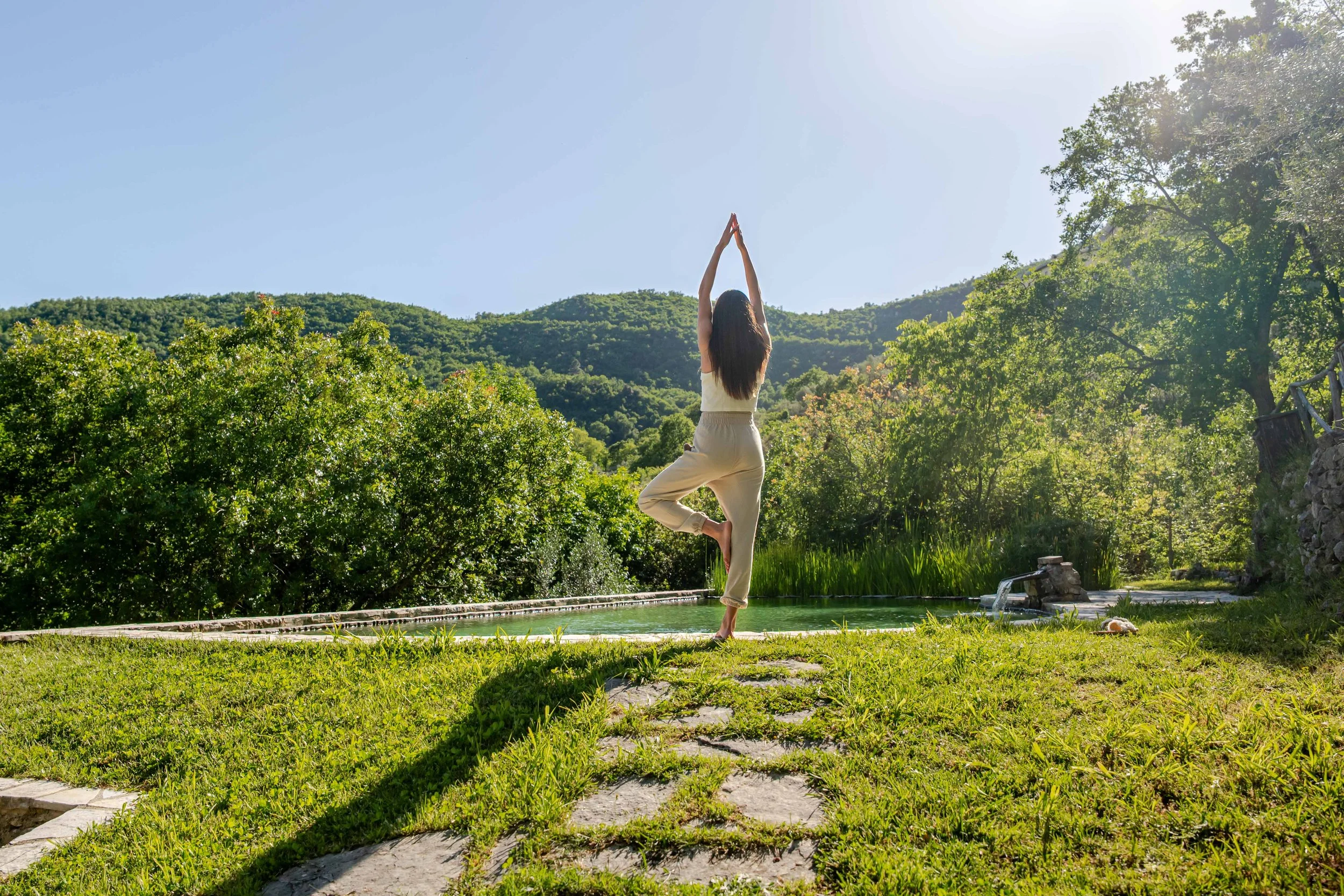Woman practicing yoga outdoors on a sunny day in a lush green setting, standing on one leg in a tree pose with arms raised above her head. Mountains and trees are in the background.