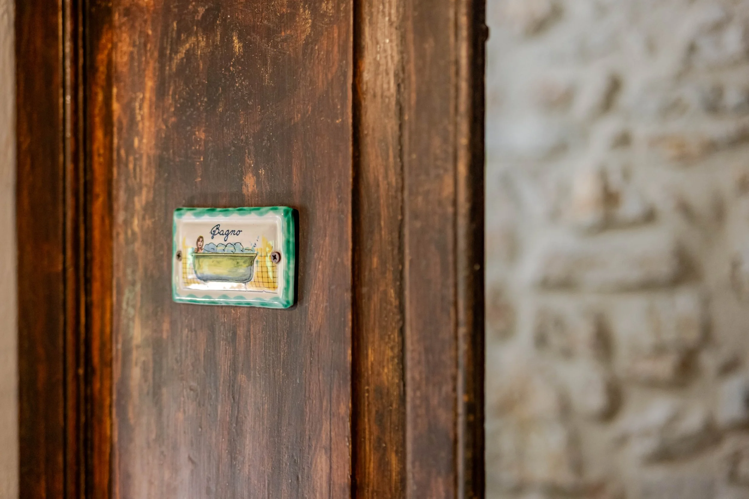 Close-up of a small ceramic sign labeled 'Bagno' with an illustration of a bathtub, mounted on a wooden wall near a stone wall.