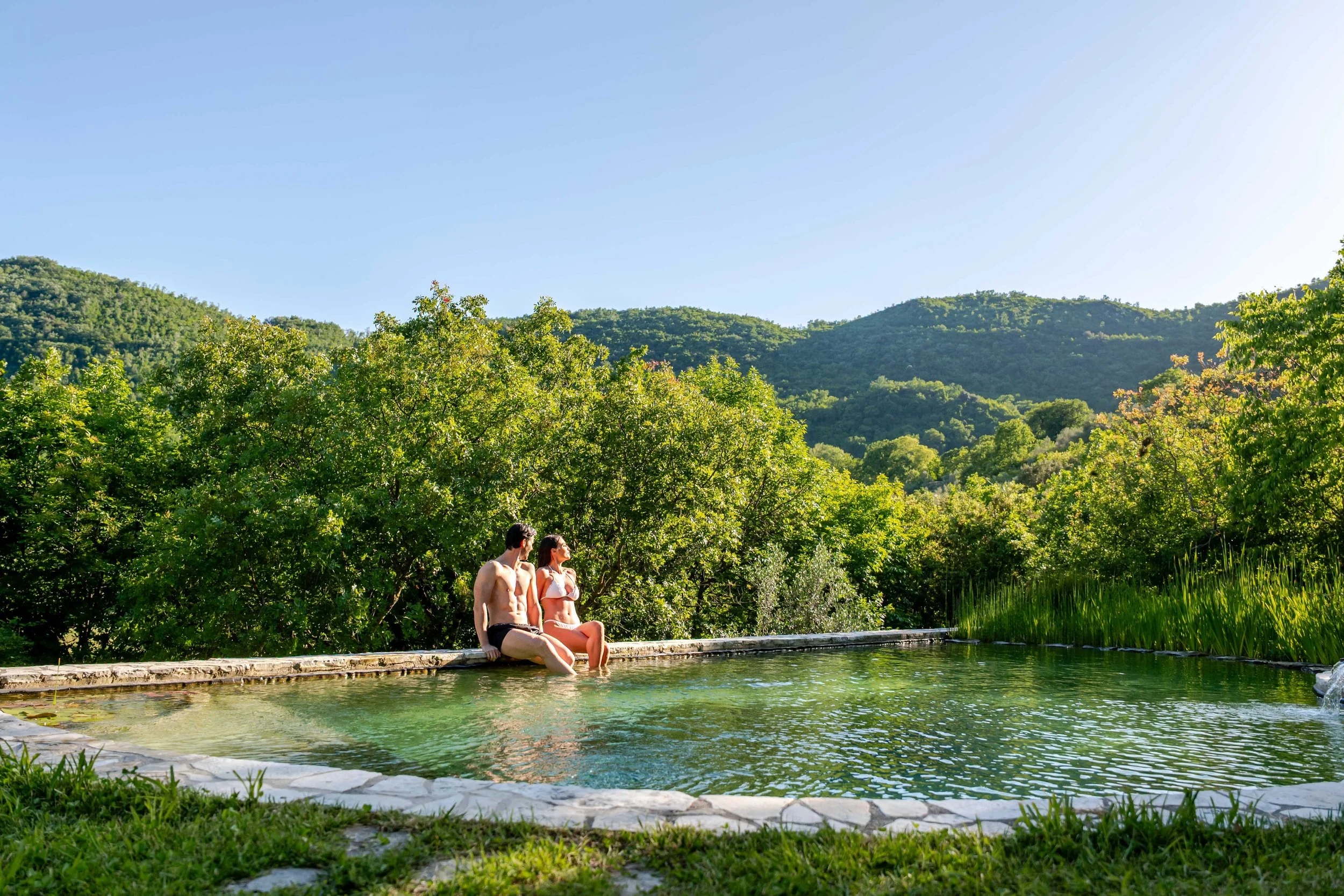 A man and woman sit at the edge of a natural hot spring surrounded by lush green trees and rolling hills on a sunny day.