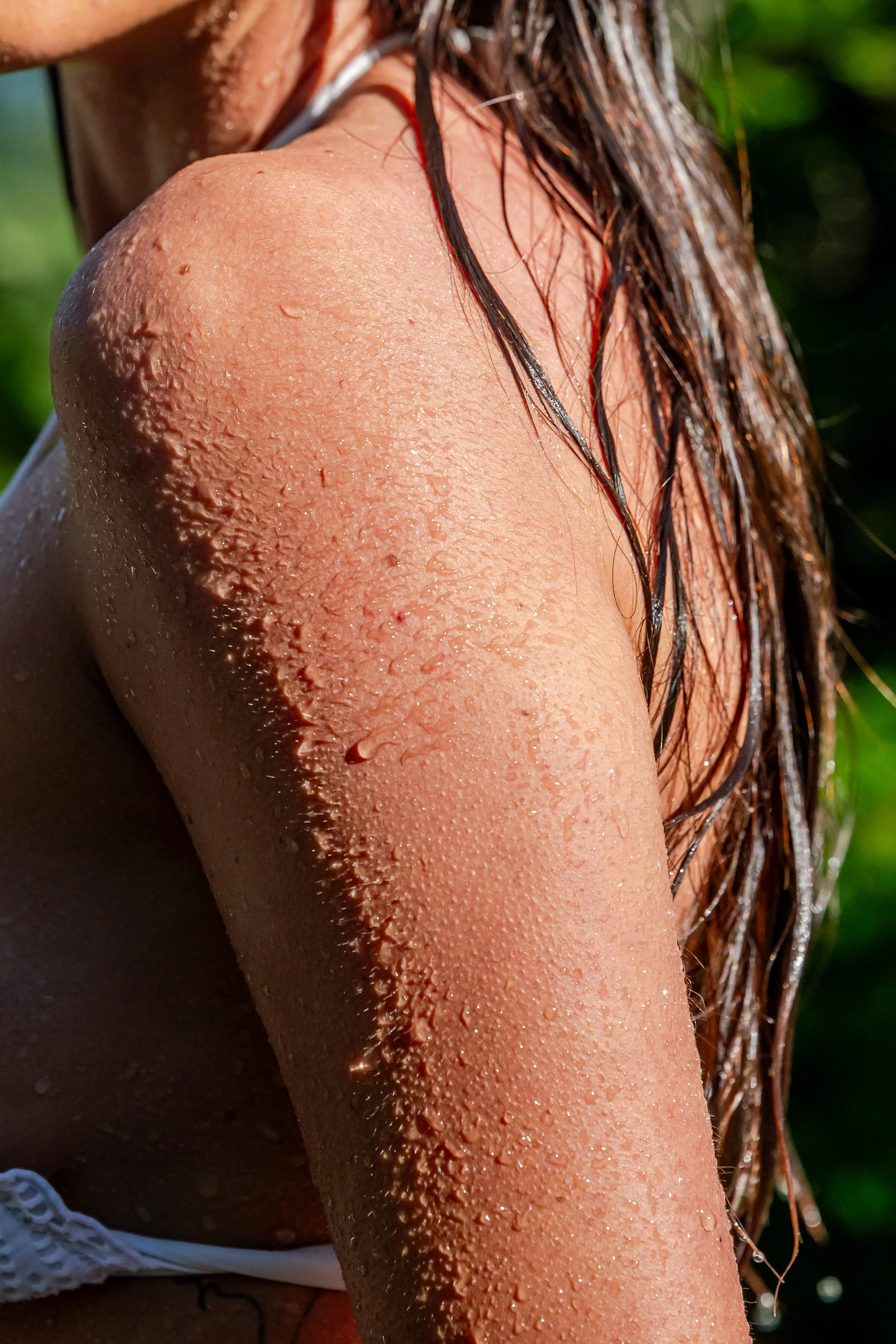 Close-up of a wet, glistening shoulder and upper arm of a person with water droplets and hair hanging down, with a blurred green background.
