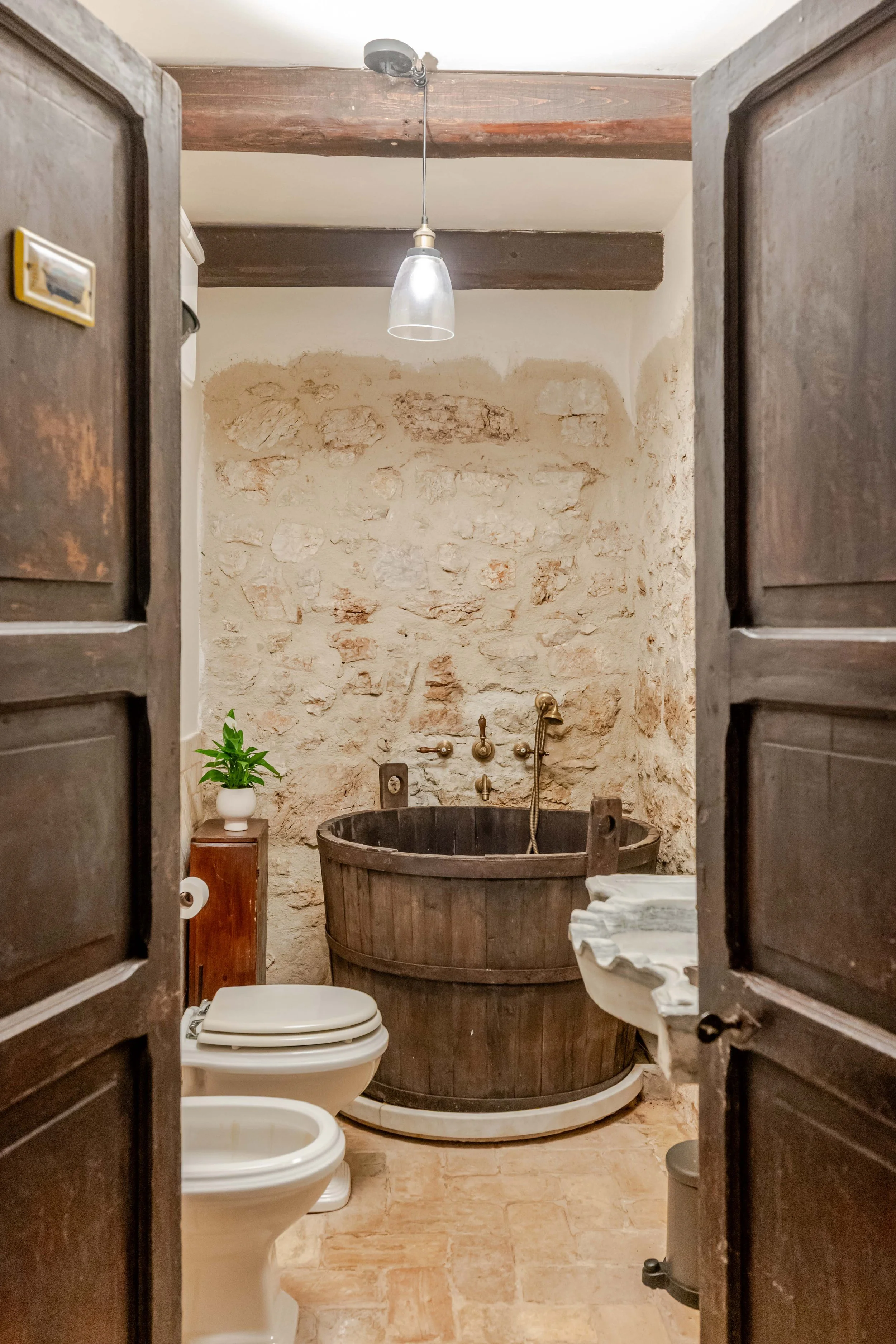 A rustic bathroom with wooden doors, a stone wall, a vintage bathtub, a toilet, a small plant, and a sink.