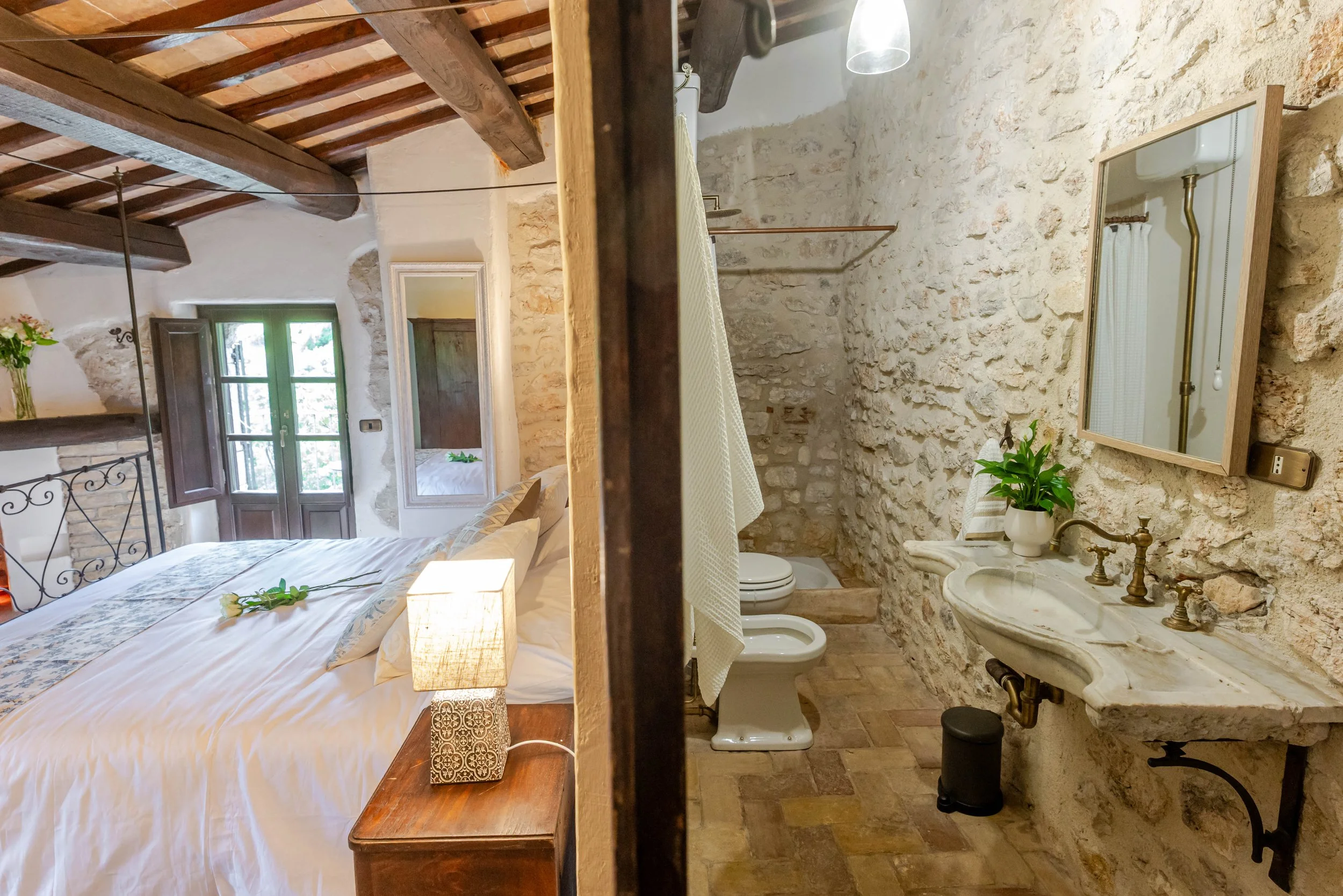 A cozy bedroom with a large bed, bedside table with a lamp, and a window with wooden shutters; adjacent bathroom with stone walls, toilet, sink with a mirror, and a shower area.