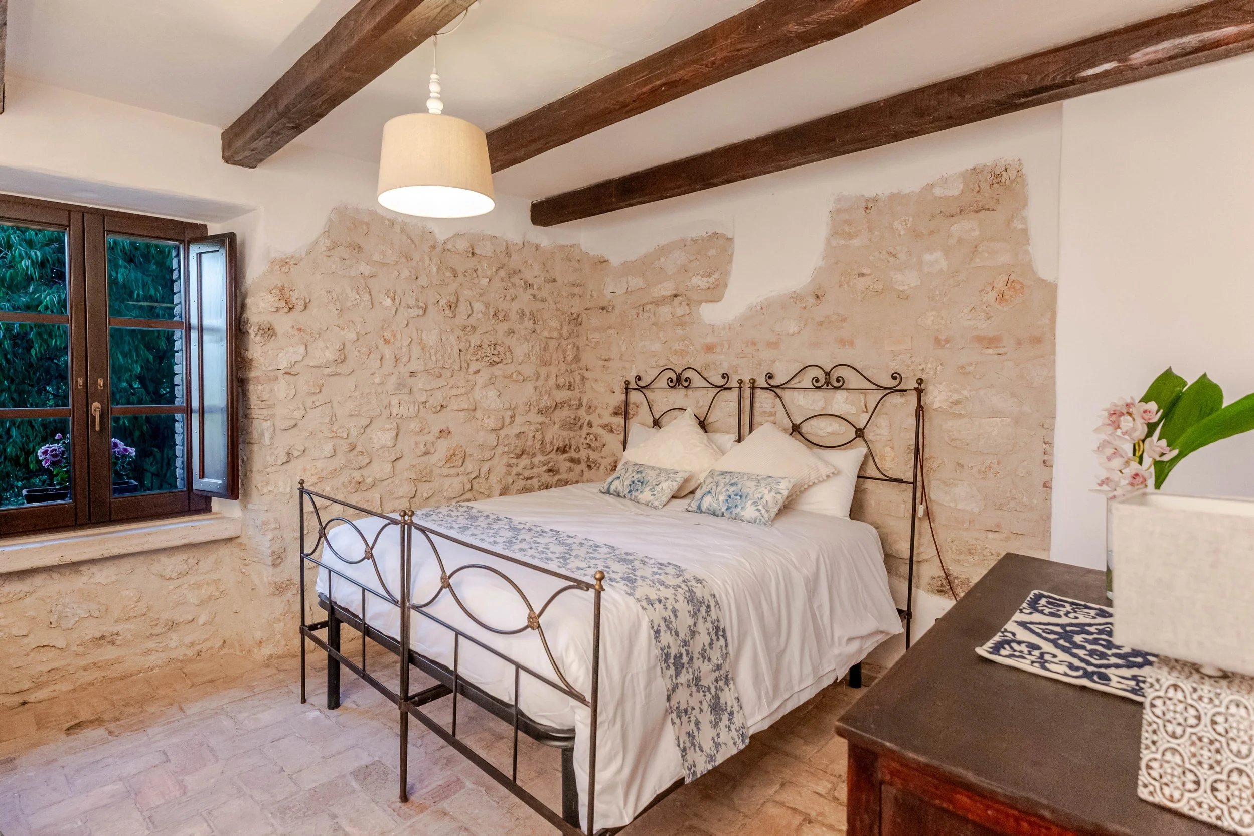 A cozy bedroom with stone walls, a black metal bed frame, white bedding, and decorative pillows. A window with open shutters shows greenery outside; a wooden dresser with a flower pot and cloths is in the corner. Exposed wood beams are on the ceiling