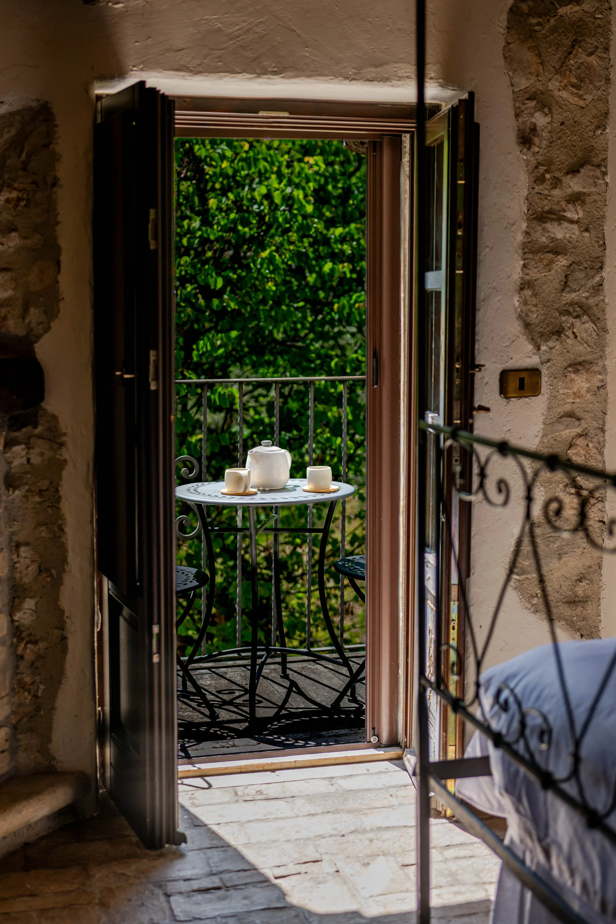 Open balcony door with a small round table and two chairs, a teapot, and two cups, overlooking green trees.
