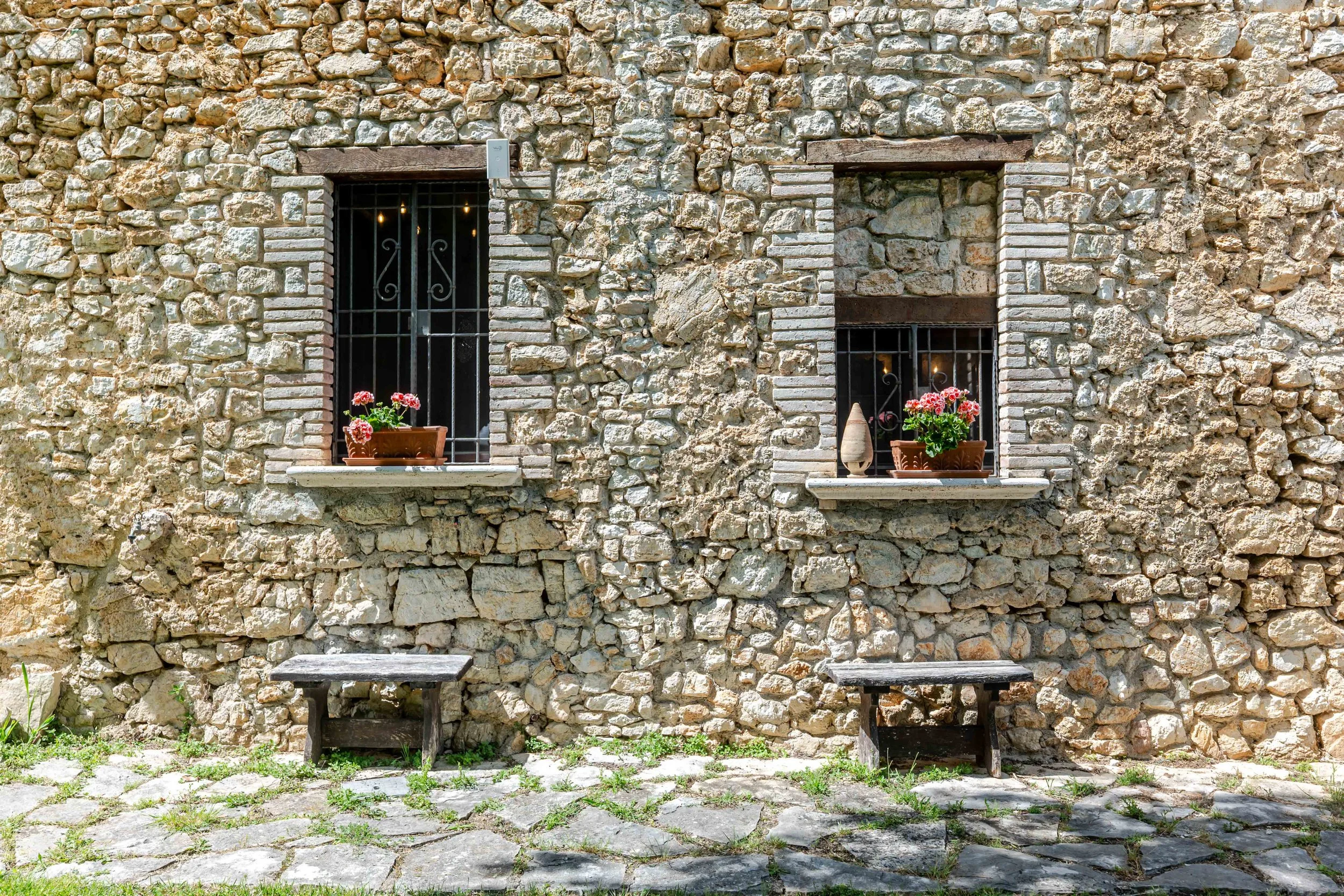 Stone wall exterior of a building with two windows decorated with flower pots and small decorative objects, two wooden benches in front on the cobblestone ground.