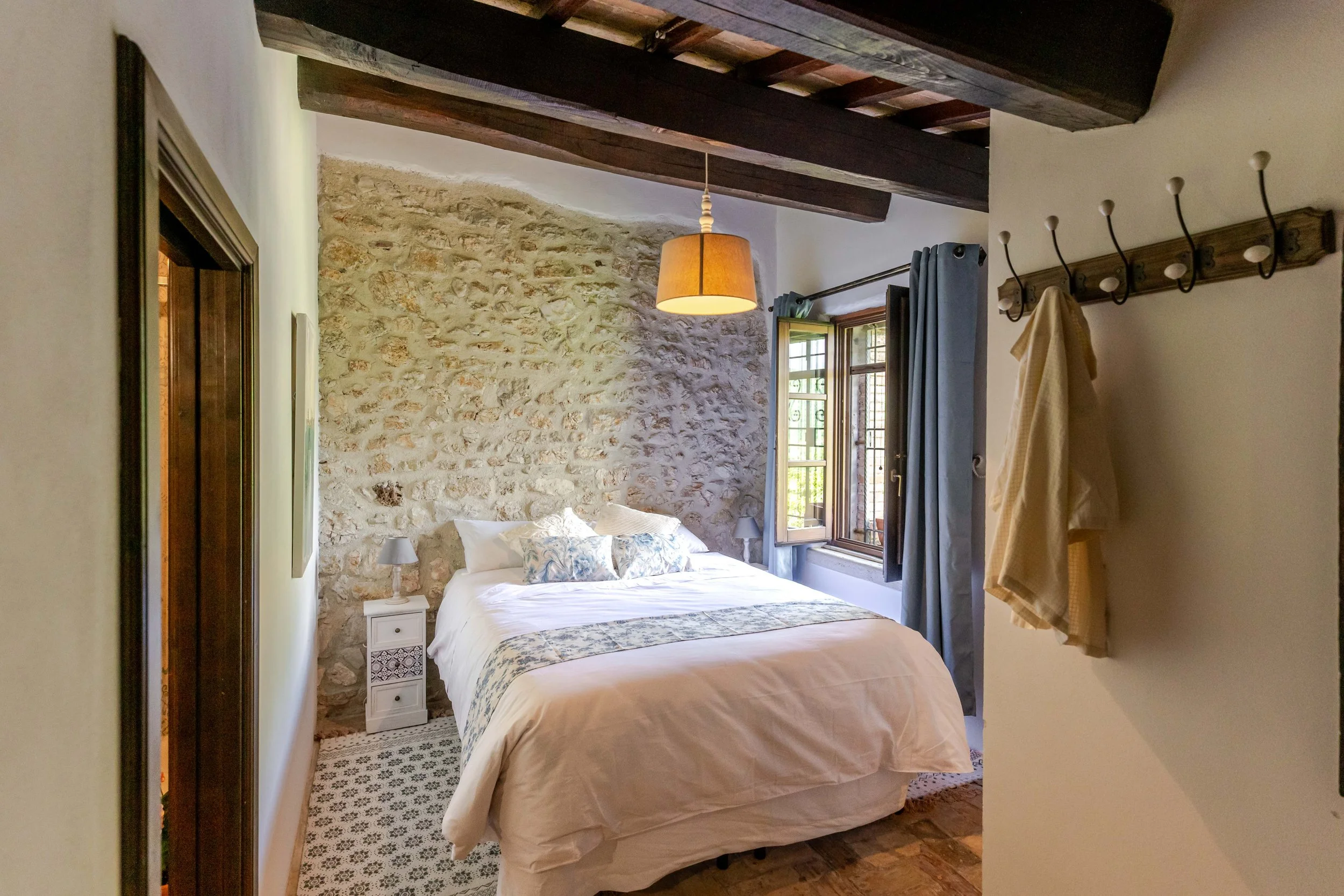 A cozy bedroom with a stone wall, a white bed with blue patterned pillows and a throw, two small nightstands with lamps, a window with blue curtains, and a wall-mounted coat rack with a yellow towel or robe hanging.